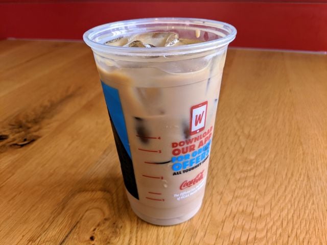 A clear plastic cup filled with iced coffee sits on a wooden table. The cup has a red “W” logo and app promotion text printed on it. Ice cubes are visible in the coffee.