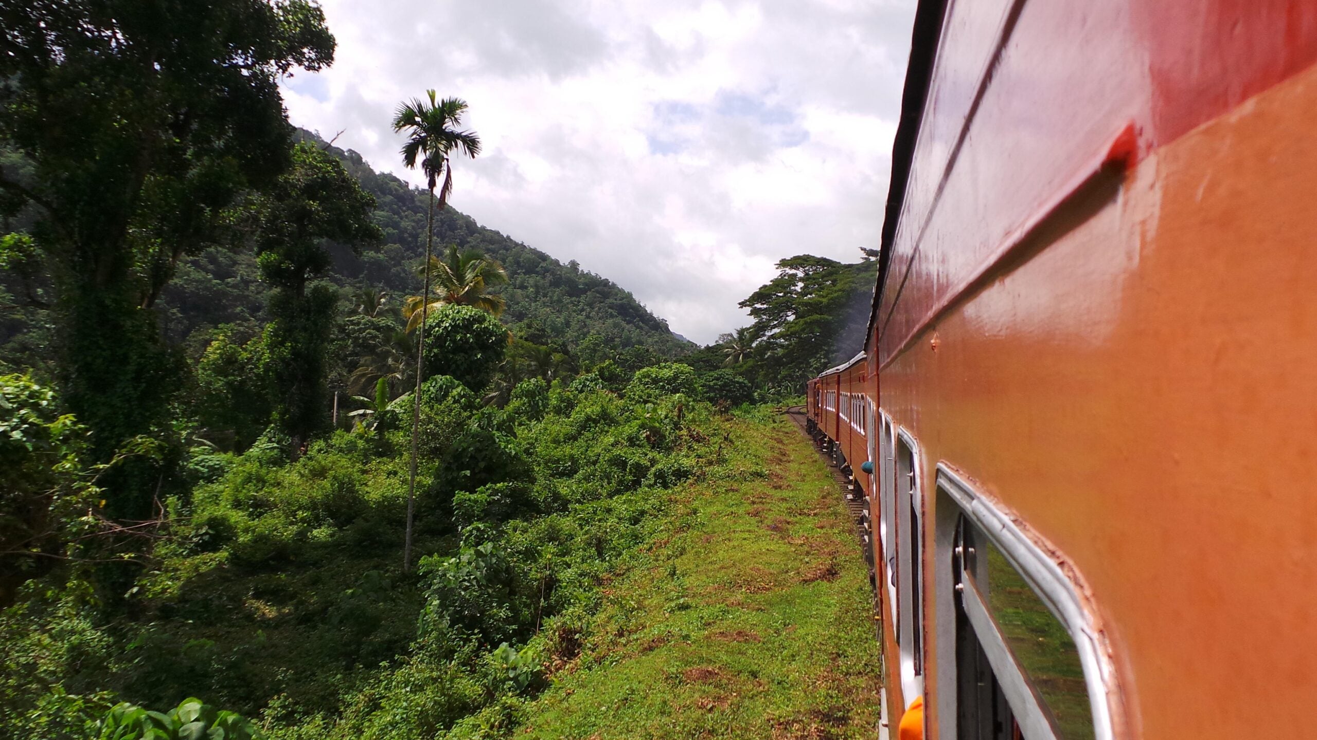 A red train travels through lush green tropical vegetation, hugging the side of a mountain under a partly cloudy sky—an iconic scene capturing the essence of Travelers’ Journeys.