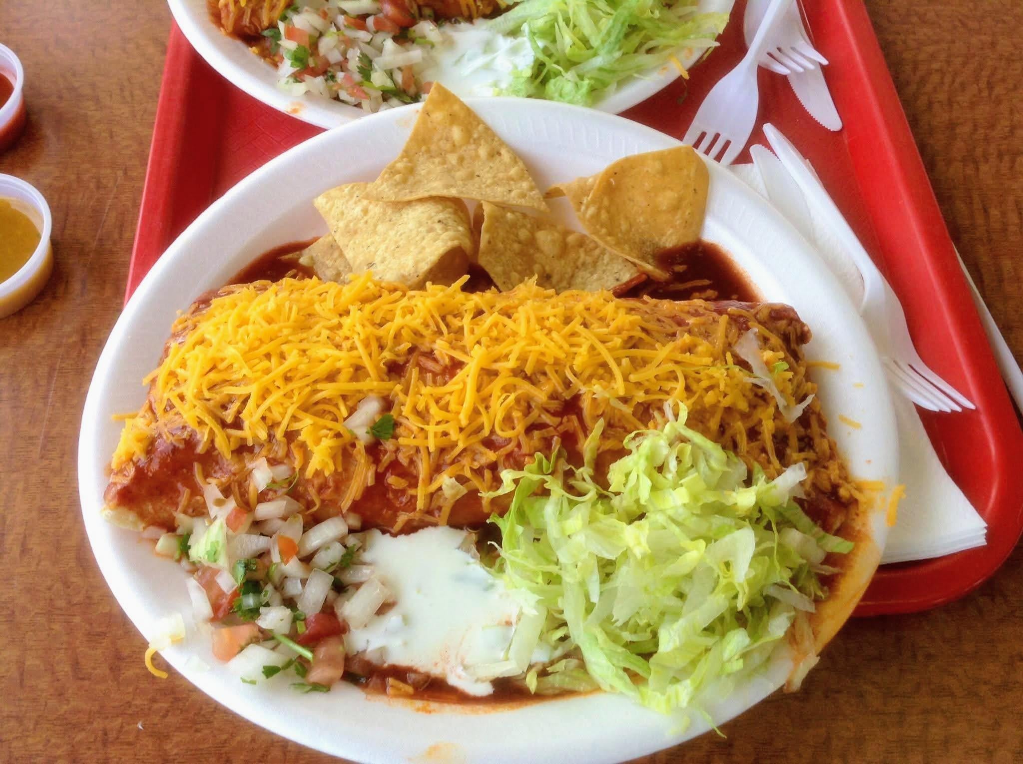A plate with a large enchilada topped with shredded cheese, lettuce, pico de gallo, tortilla chips, and sauce on a red tray. Another similar plate and plastic utensils are partially visible in the background.