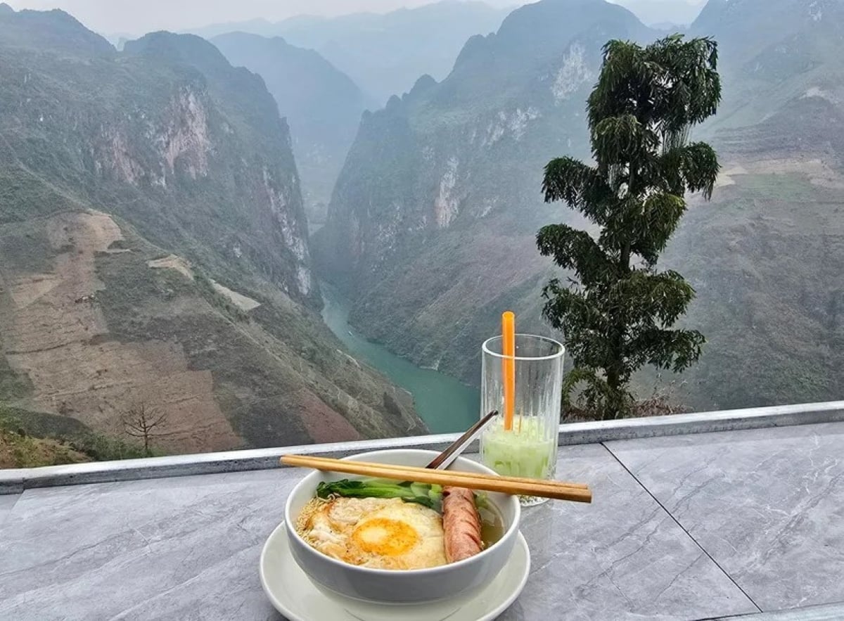 A bowl of noodles with egg and sausage, chopsticks on top, and a cold drink sit on a table overlooking a river winding through green mountain cliffs under a cloudy sky—a perfect example of the amazing food people ate while on vacation.