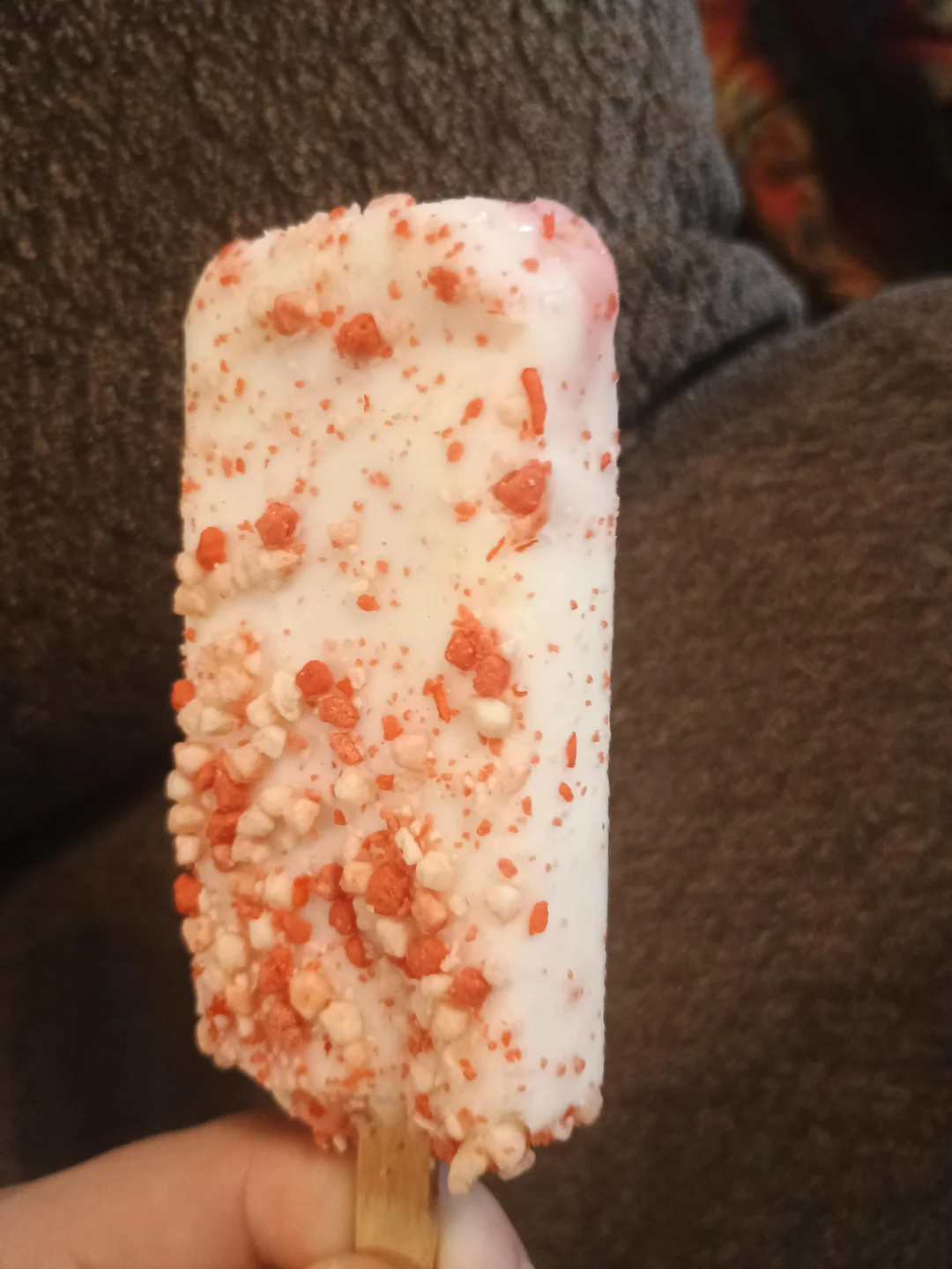A hand holds a rectangular ice cream bar coated in white icing and covered with red and white crunchy crumbs, against a brown textured background.