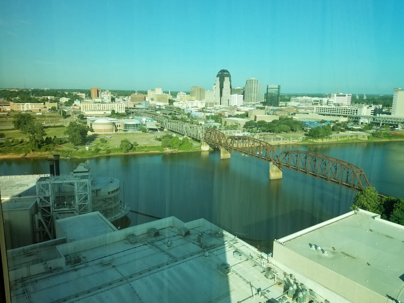 View of a city skyline with tall buildings, a rusted railway bridge crossing a river, and various structures along the riverbank under a clear blue sky. Photo is taken from a high vantage point.