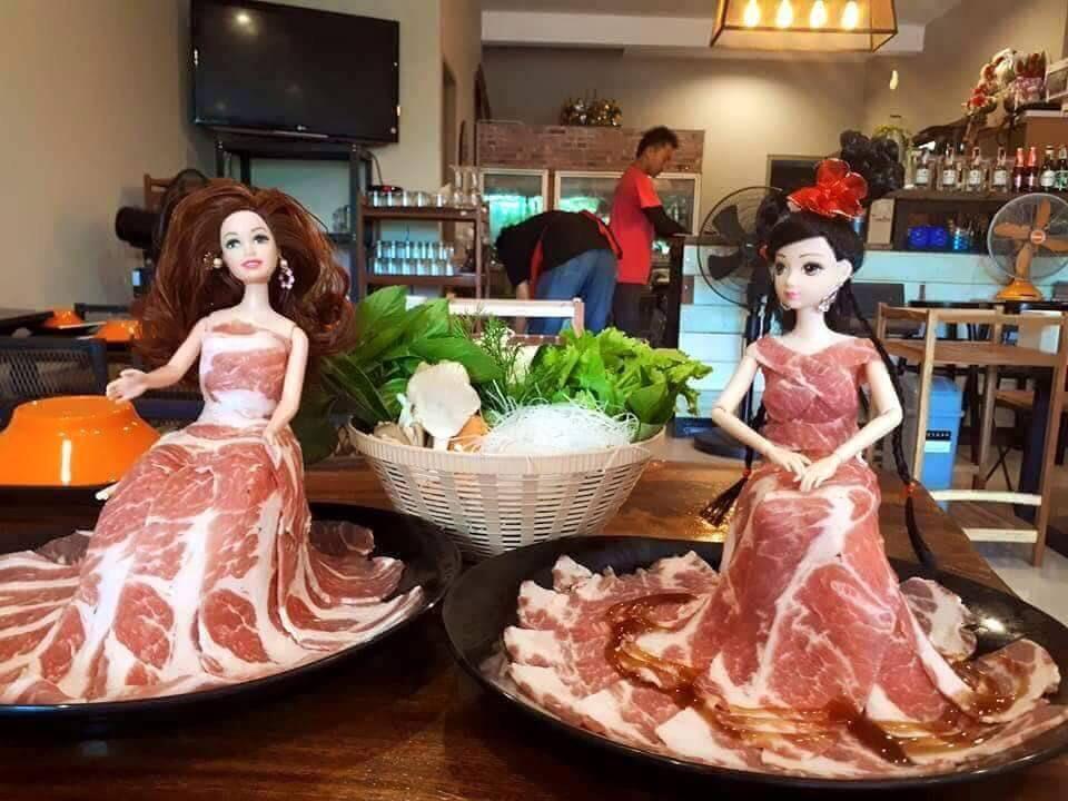 Two dolls are seated on plates, each wearing a dress made of raw meat slices arranged in a skirt shape, with a basket of vegetables and herbs placed between them on a dining table in a restaurant setting.