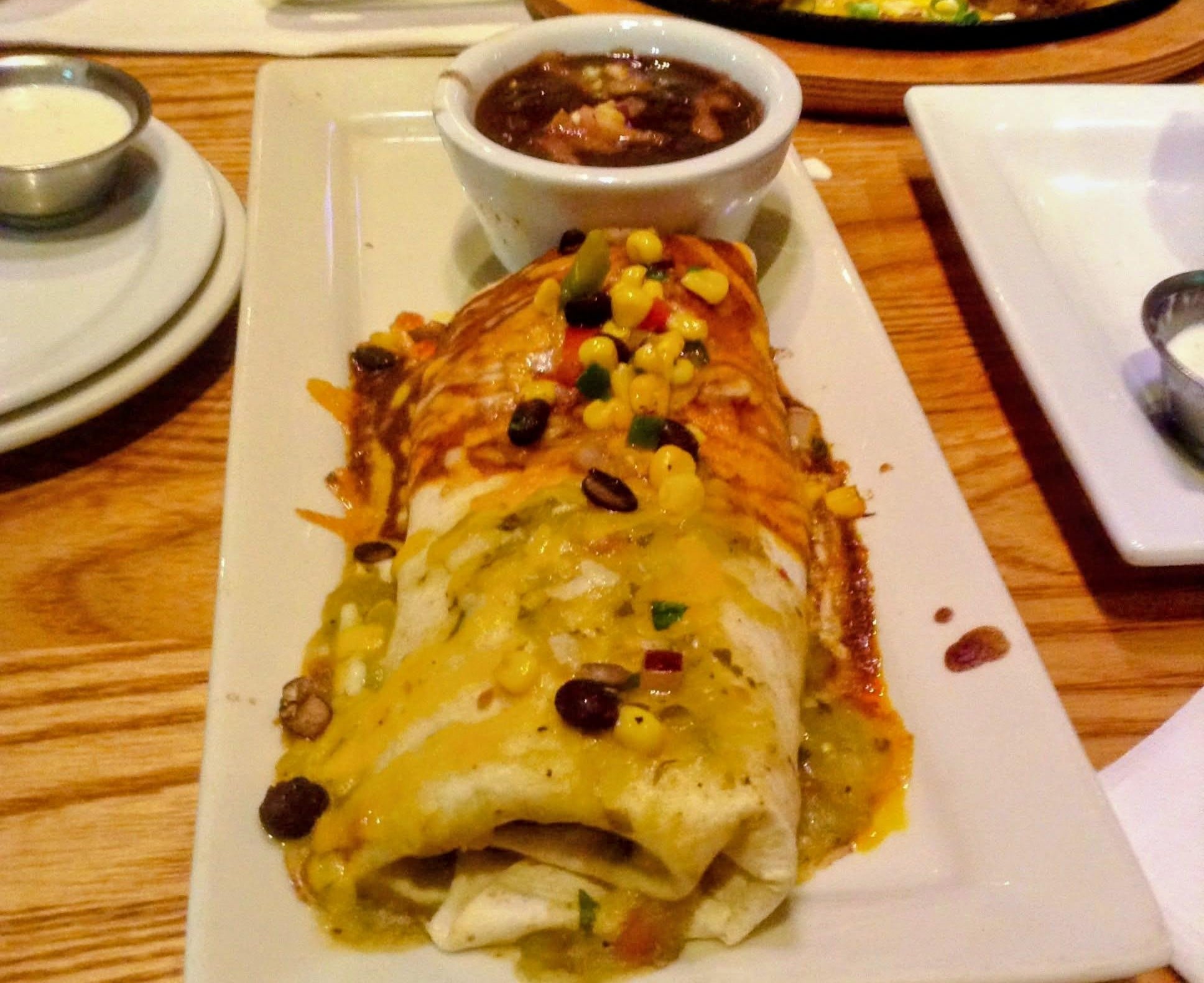 A burrito topped with melted cheese, corn, and beans sits on a rectangular plate, next to a small bowl of black bean soup.