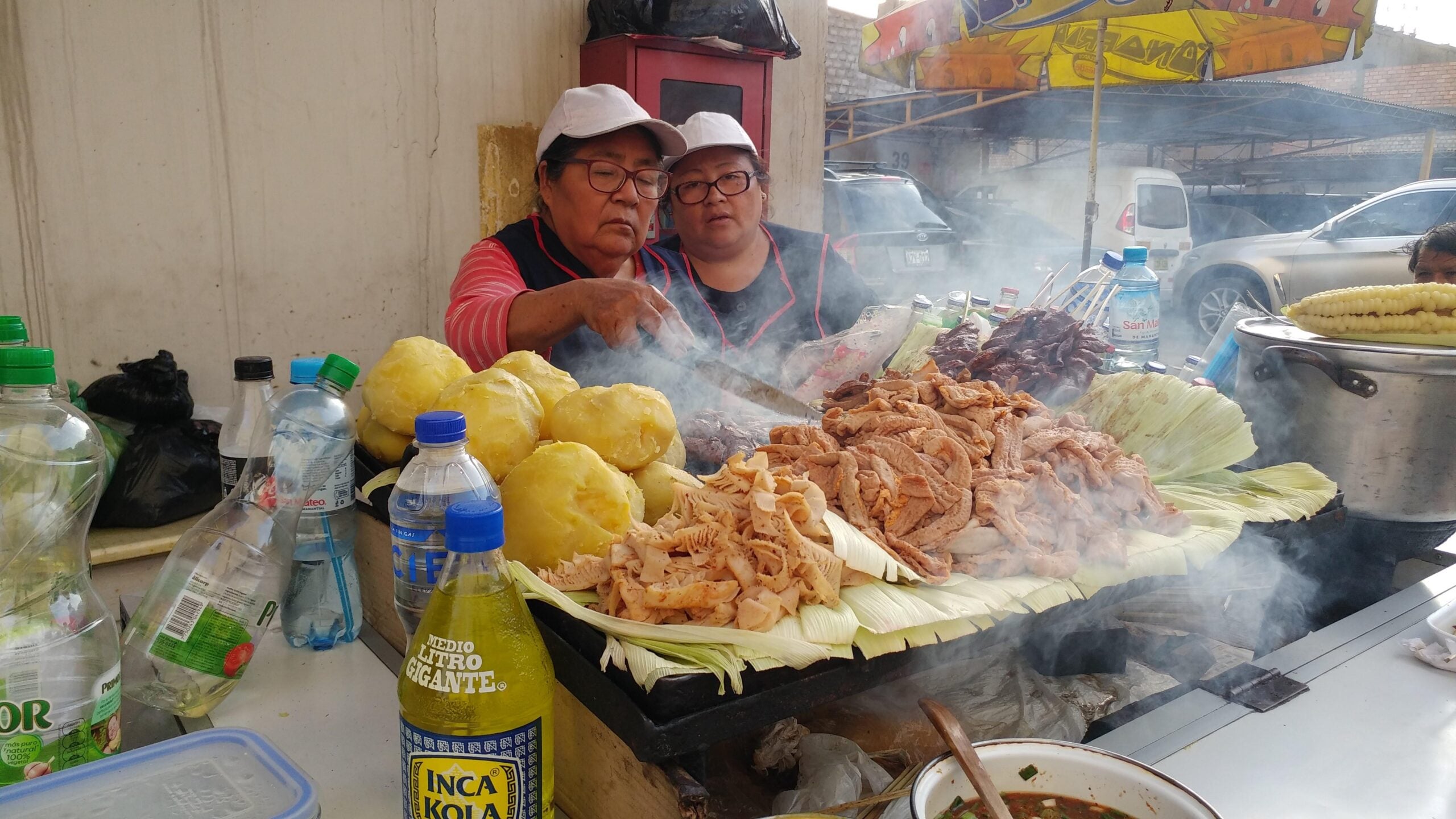 Two women wearing white hats cook meat and other food on a smoky street food grill, surrounded by bottles and large piles of yellow potatoes—truly the kind of amazing food people ate while on vacation. A bowl and drink bottles are visible in the foreground, capturing the vibrant energy of this authentic culinary experience.