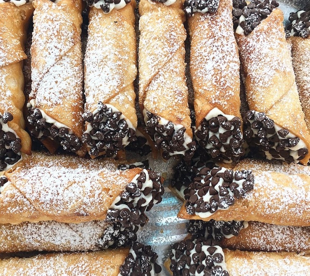 Several homemade cannolis filled with creamy filling and dipped in chocolate chips are arranged closely together. The golden shells are dusted with powdered sugar.