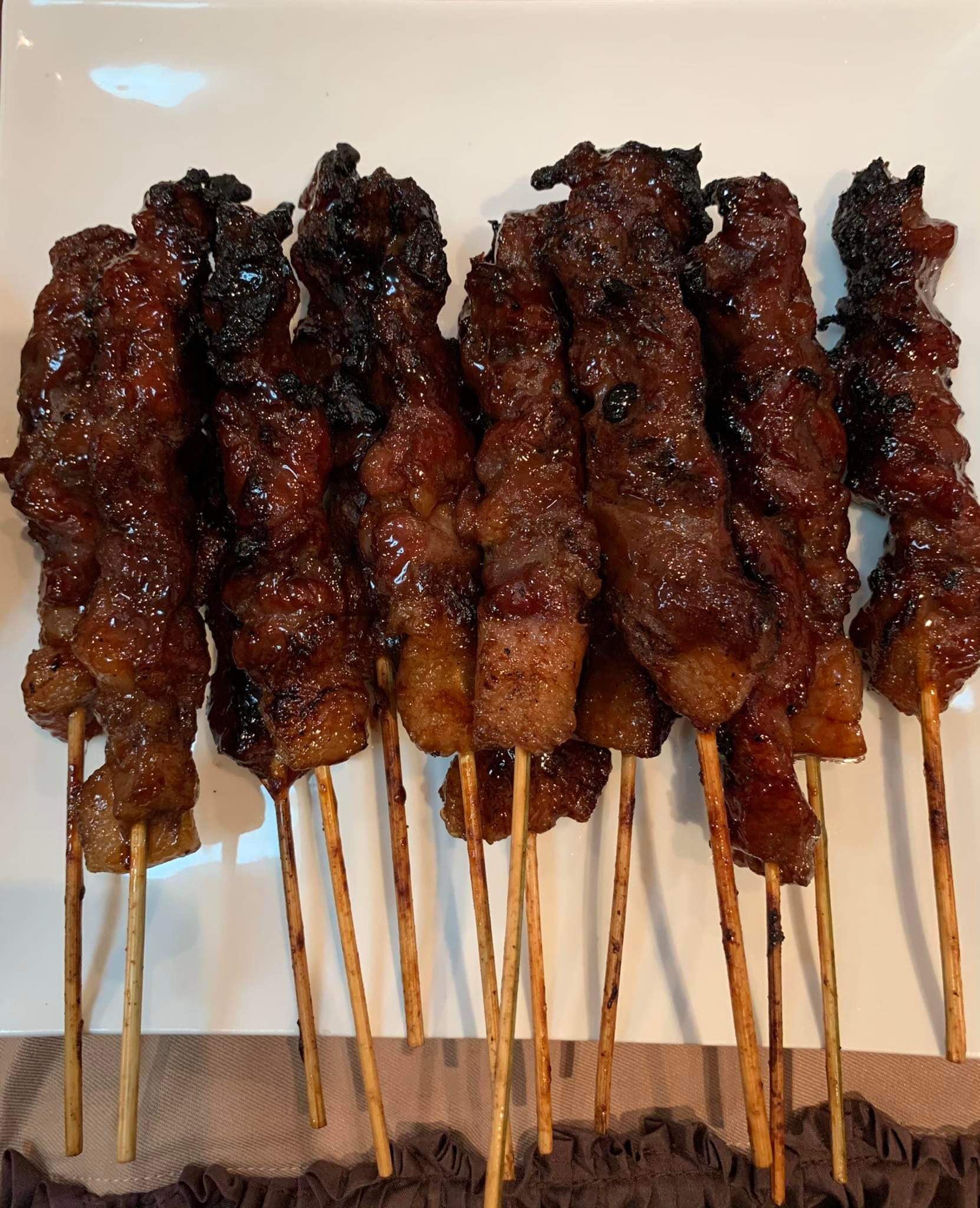 Seven skewers of grilled meat, glazed with a shiny, caramelized sauce, are arranged side by side on a white plate. The edges of the meat appear slightly charred.
