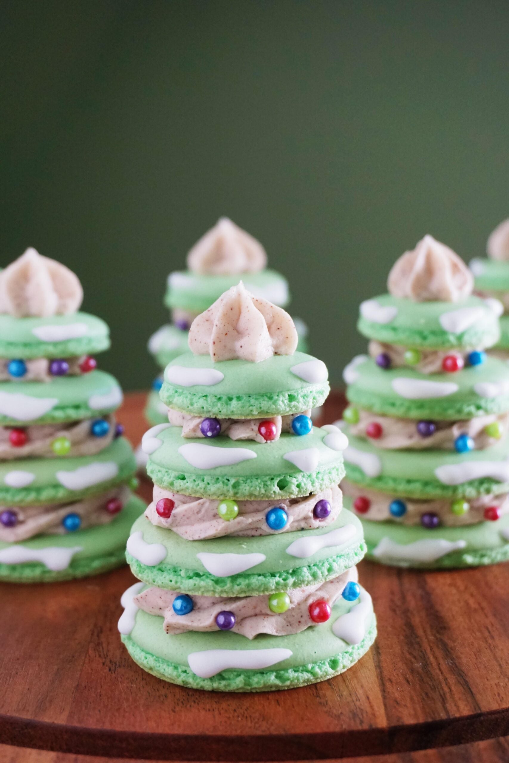 Green macaron cookies stacked and decorated with white icing and colorful round sprinkles to resemble Christmas trees, topped with a swirl of frosting, are displayed on a wooden surface against a green background—perfect homemade Christmas treats for the holiday season.