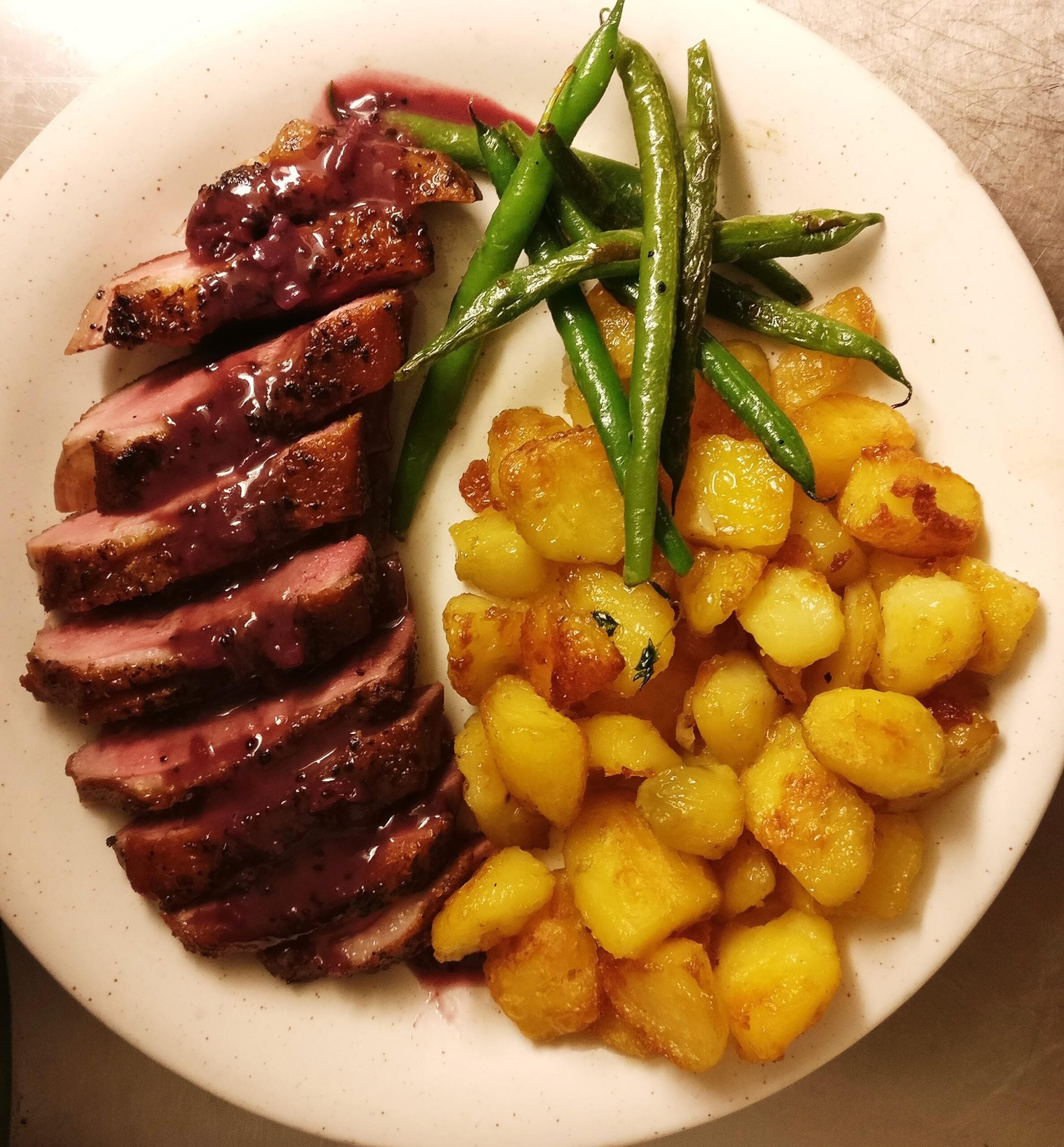 Sliced duck breast with a dark berry sauce, crispy roasted potatoes, and sautéed green beans arranged neatly on a white plate.