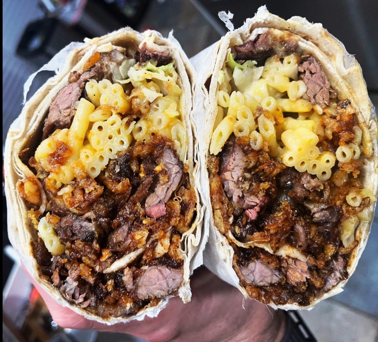 A close-up of a burrito cut in half, revealing fillings of macaroni and cheese, grilled beef, and crispy brown bits, all wrapped in a tortilla.