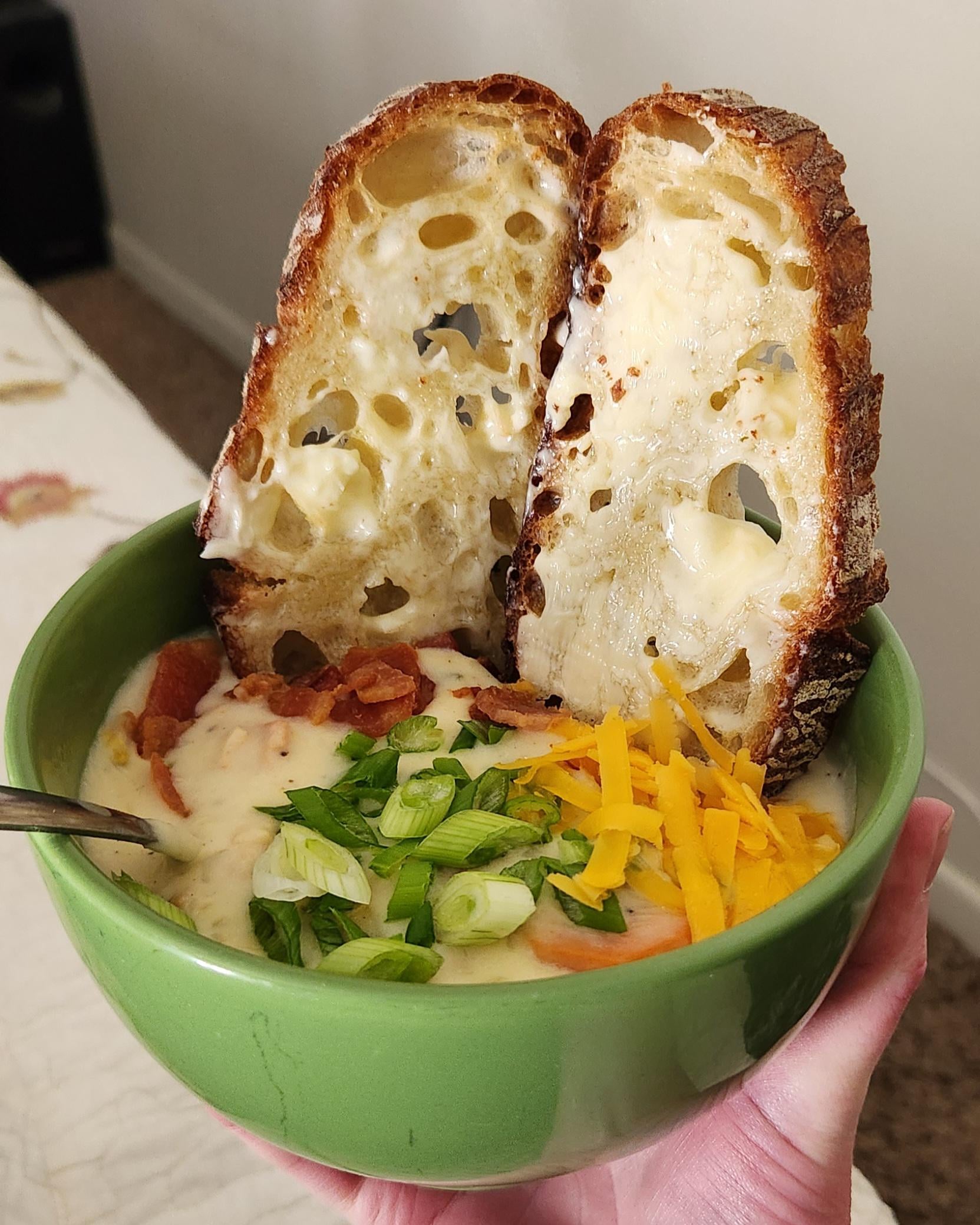 A green bowl of creamy soup topped with chopped green onions, shredded cheddar cheese, and bacon bits, with two slices of buttered, toasted bread sticking up from the bowl.