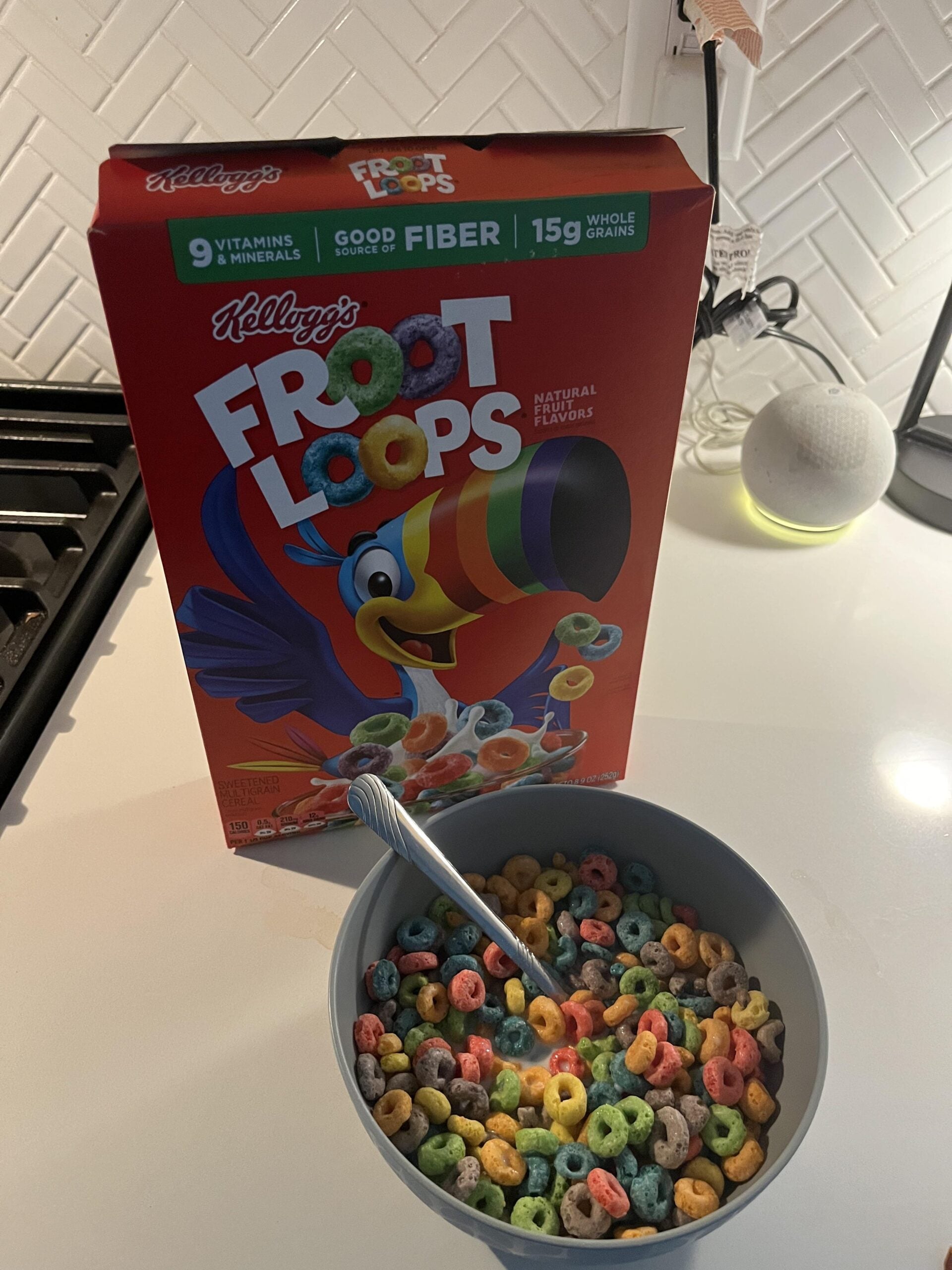 A box of Froot Loops cereal stands behind a bowl filled with Froot Loops and milk on a kitchen counter. A spoon is in the bowl, and part of a stovetop and a smart speaker are visible nearby.