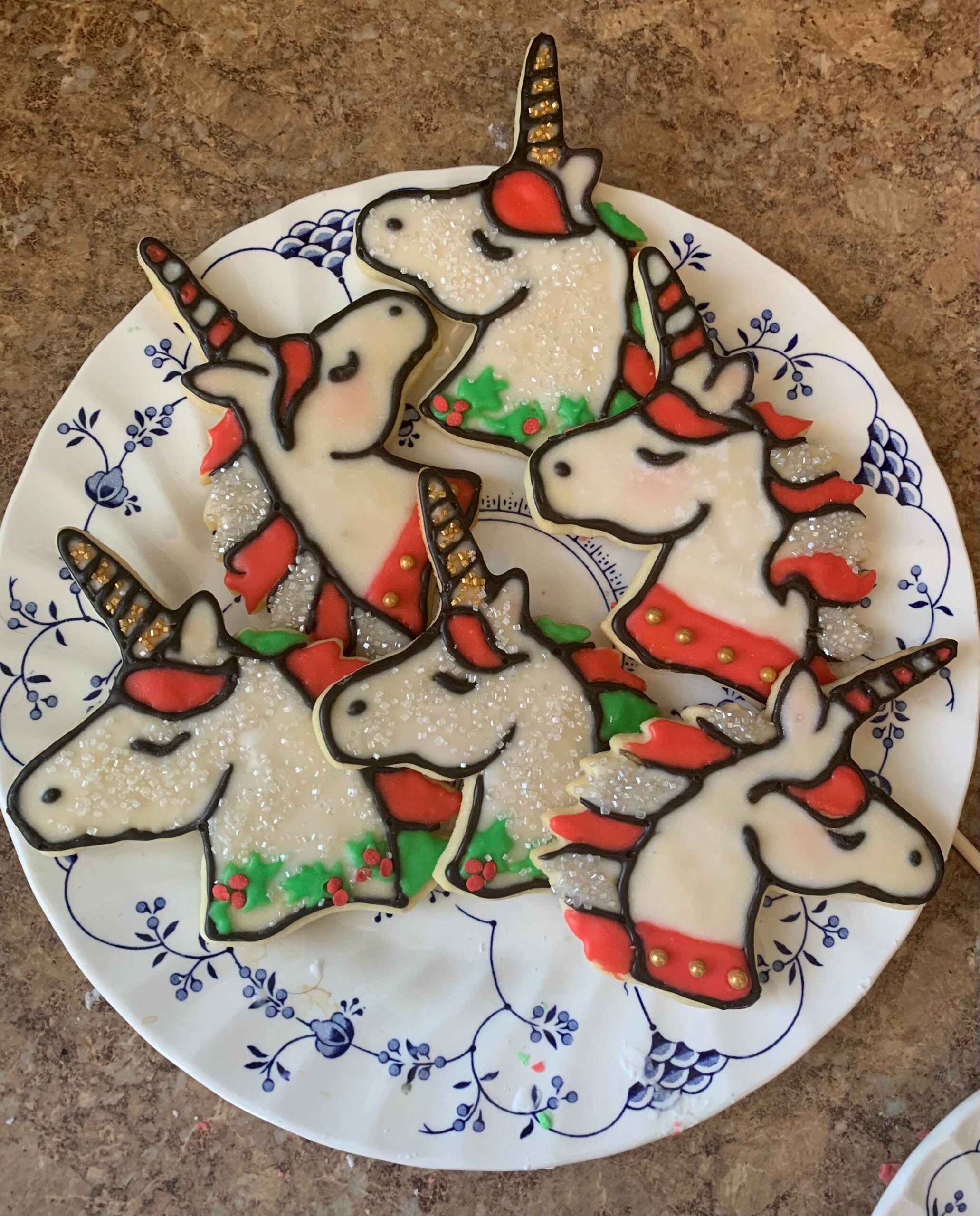 A plate with seven decorated unicorn cookies, featuring white icing, red and green accents, gold horns, and black outlines, sits on a brown countertop—perfect homemade Christmas treats to brighten any holiday gathering.