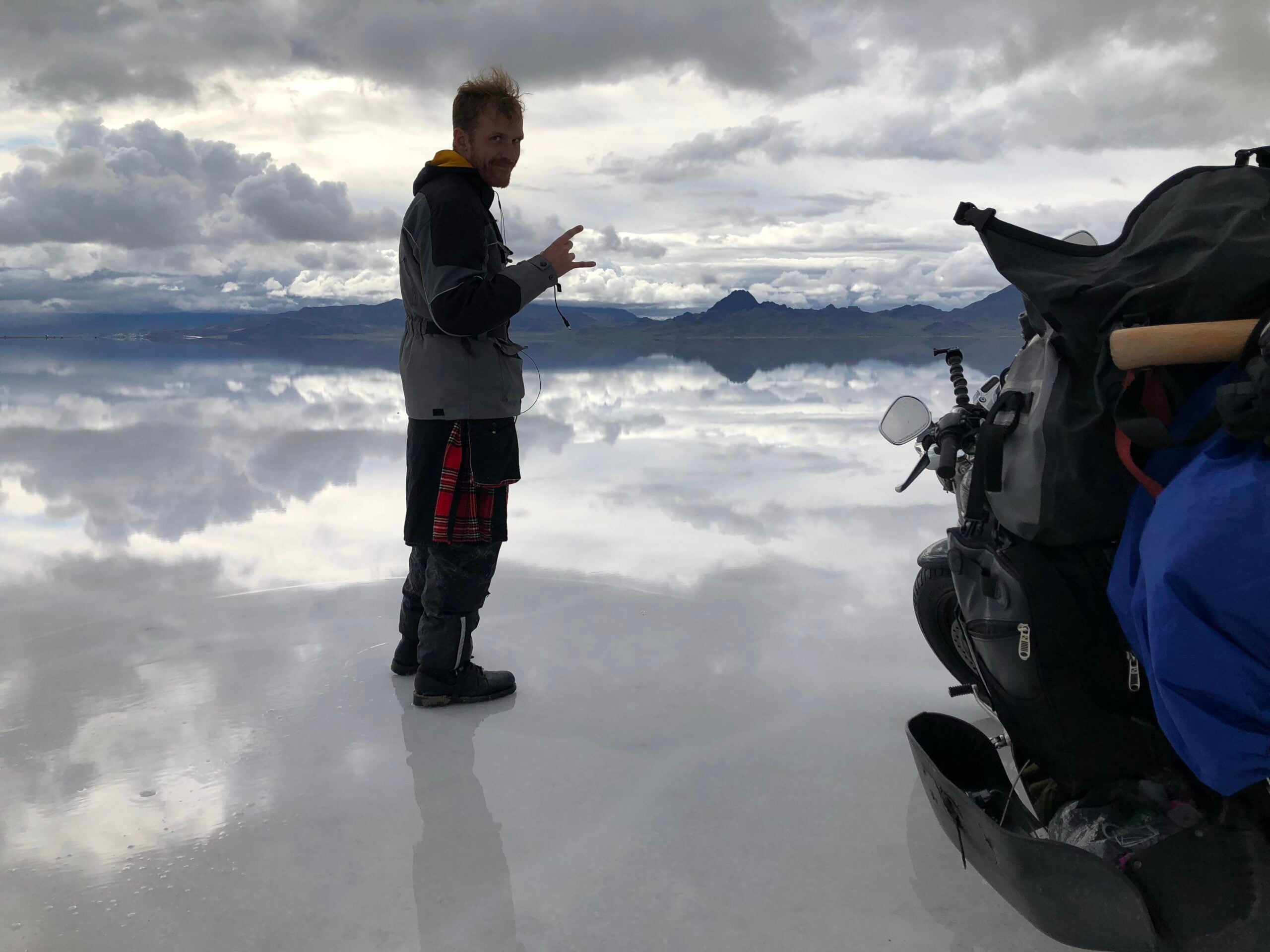 A person stands on a reflective salt flat near a motorcycle, dressed in boots, a jacket, and a kilt—ready for road trips—with mountains and a cloudy sky forming the dramatic backdrop.