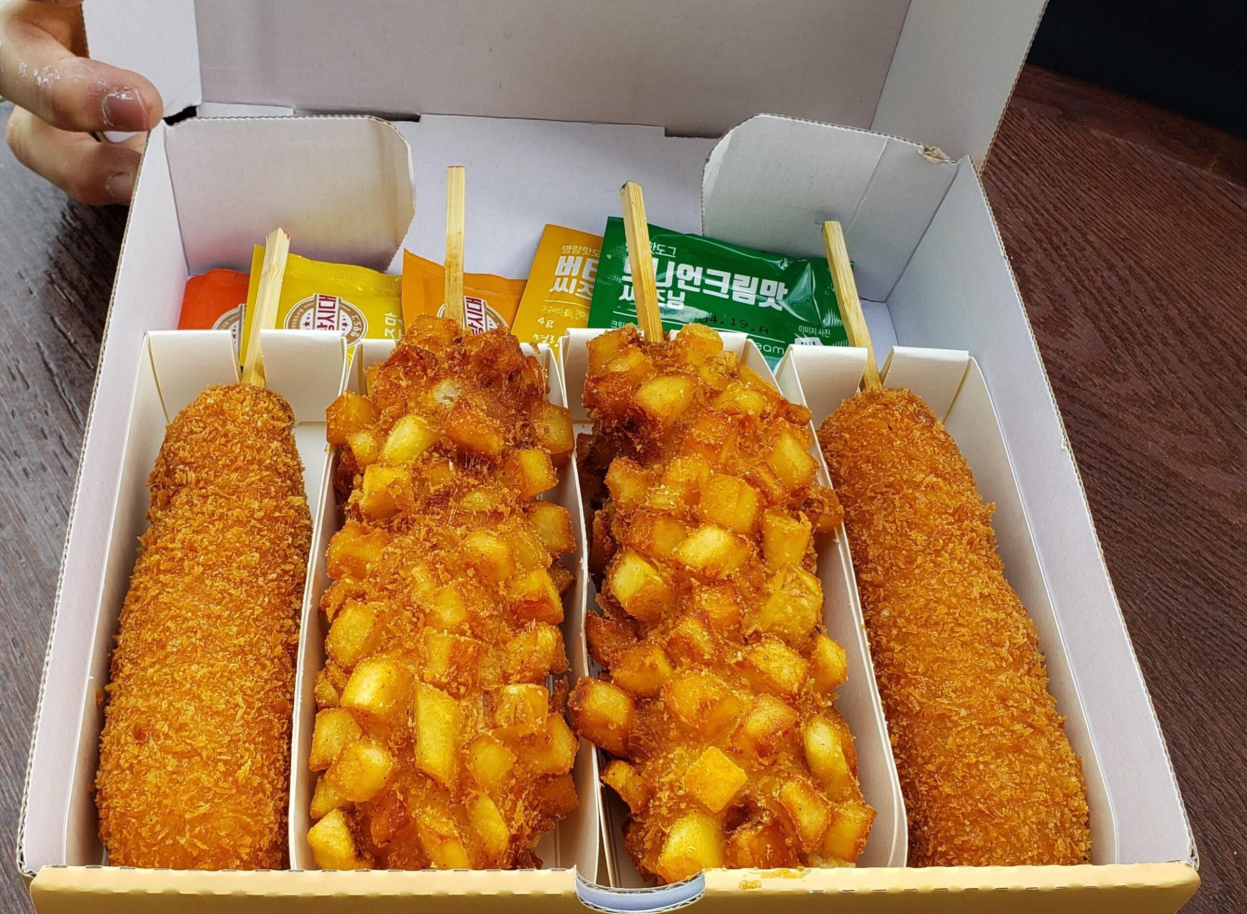 A box contains four Korean corn dogs: two with a plain fried coating and two coated with crispy cubed potatoes. Packets of mustard and sauce are included in the back of the box. A hand is visible in the corner.