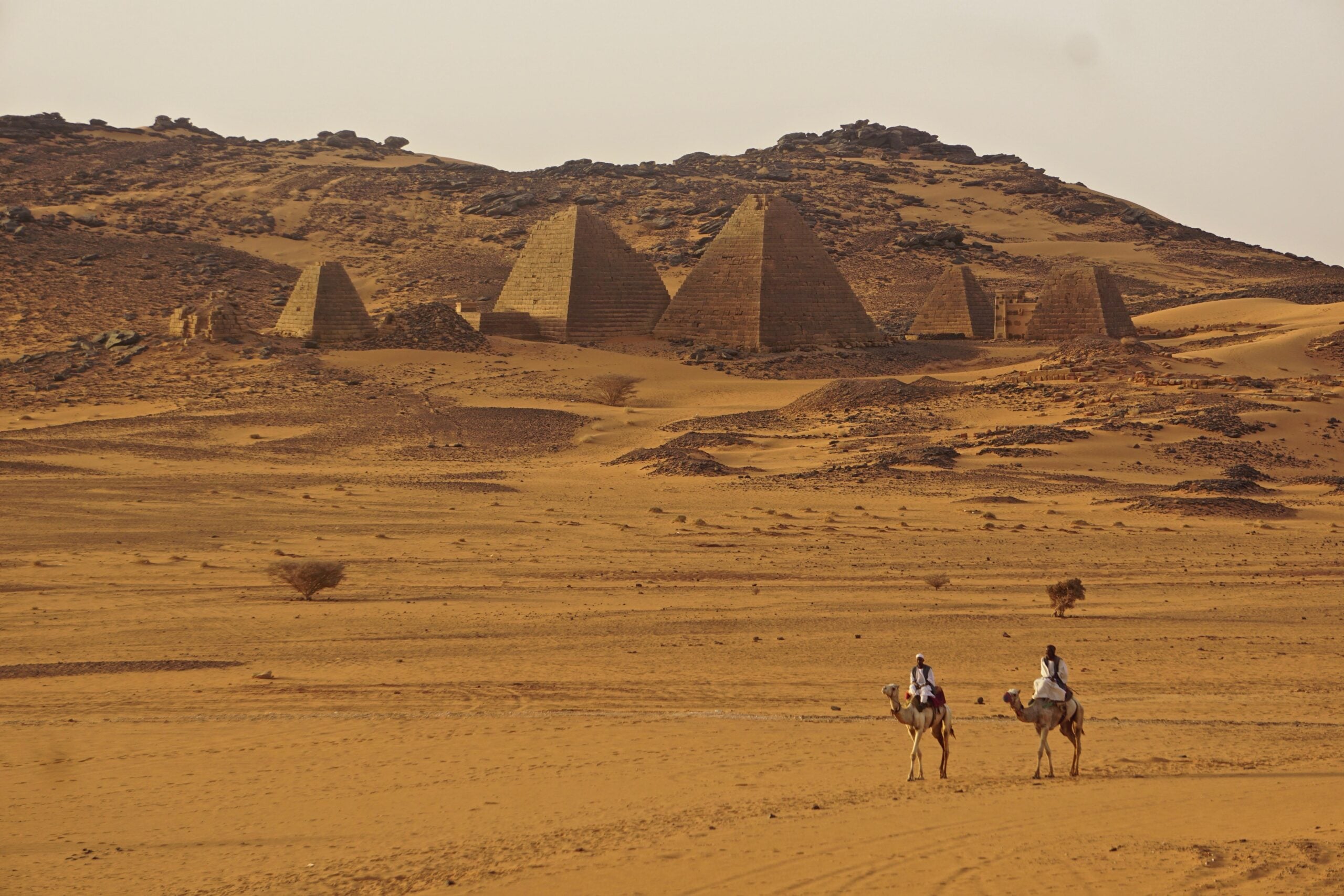 Two people ride camels across a sandy desert toward ancient pyramids, partially buried and surrounded by rocky hills under a clear sky.