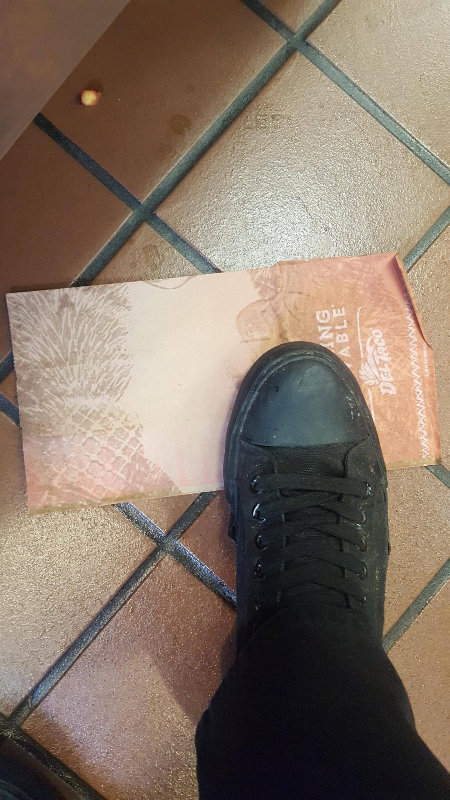 A person wearing a black shoe steps on a brown paper napkin on a tiled floor. There is a small piece of food nearby.