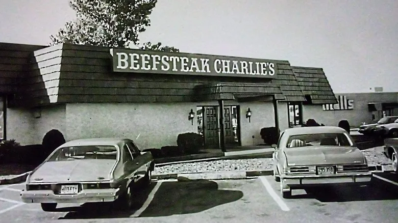 Black-and-white photo of two vintage cars parked in front of a Beefsteak Charlie’s restaurant with a large sign above the entrance.
