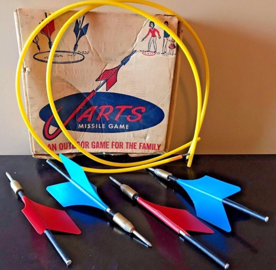 Vintage lawn darts set with three red and two blue darts, two yellow plastic rings, and a cardboard box labeled “Jarts Missile Game: An outdoor game for the family,” displayed on a dark surface.