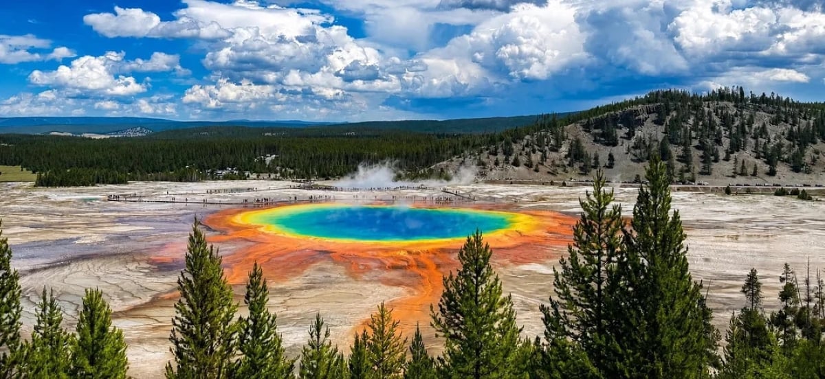 A vibrant hot spring with blue, green, yellow, and orange rings sits in a rocky area surrounded by pine trees and hills under a partly cloudy sky.