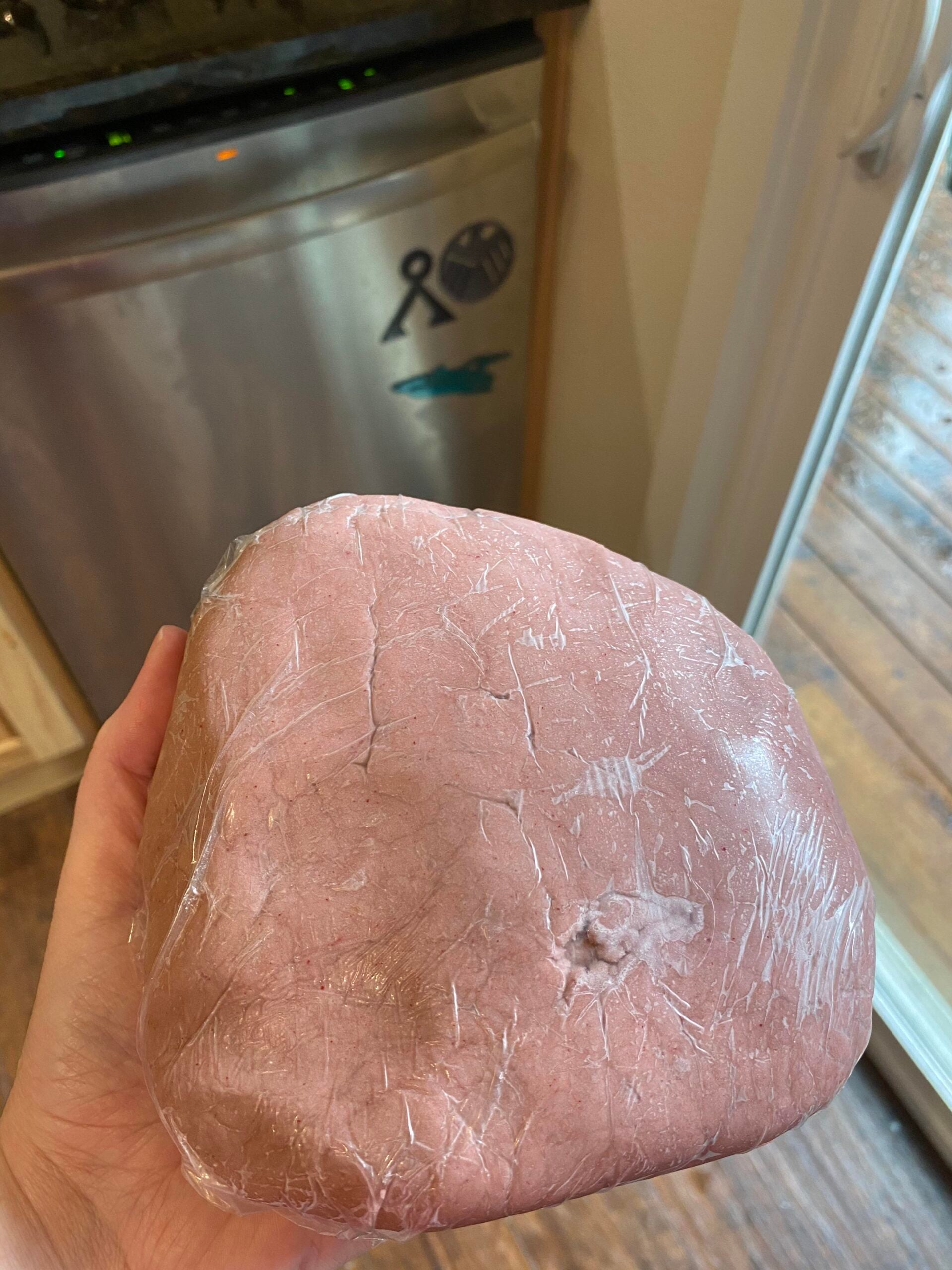 A hand holds a large chunk of pink-colored dough or clay wrapped in plastic wrap, with kitchen appliances and a window in the background.