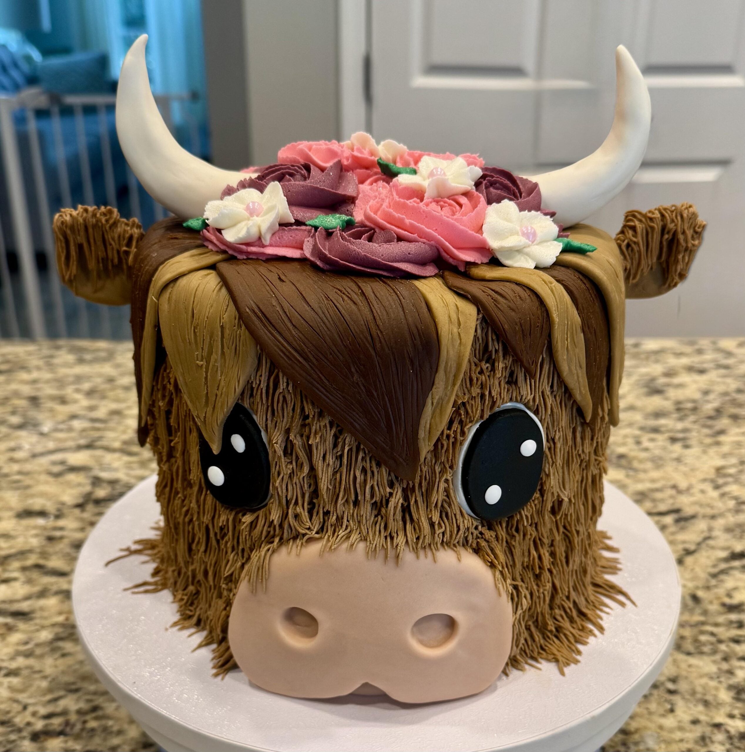 A cake decorated to look like a cute highland cow with large horns, big eyes, shaggy brown fur, and a crown of pink, purple, and white flowers on its head.