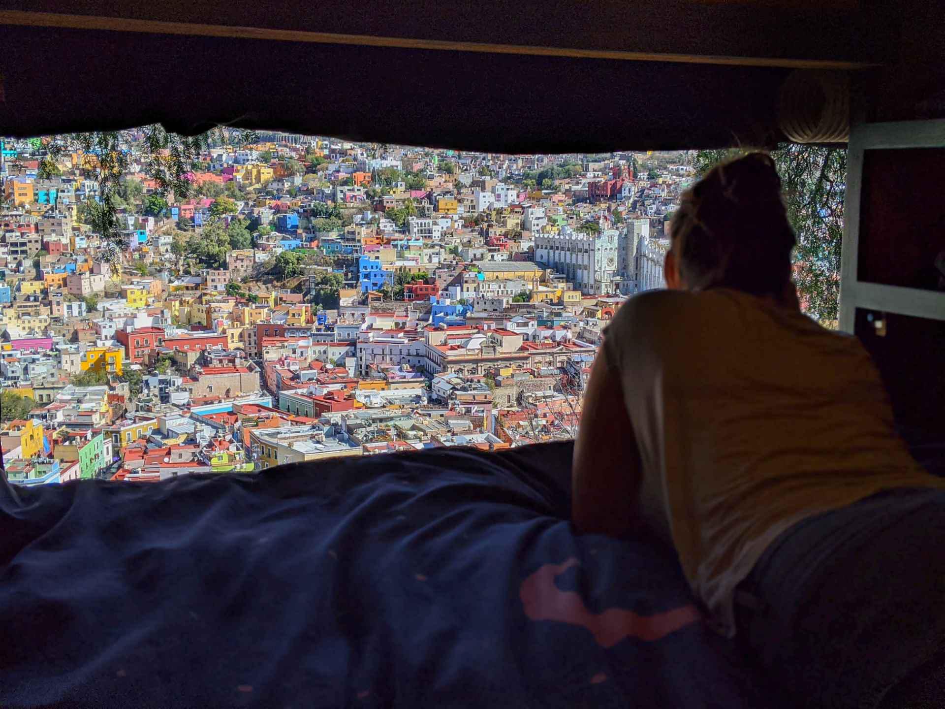 A person lies on a bed inside a dark room, looking out through a large window at a colorful, densely packed cityscape in daylight.