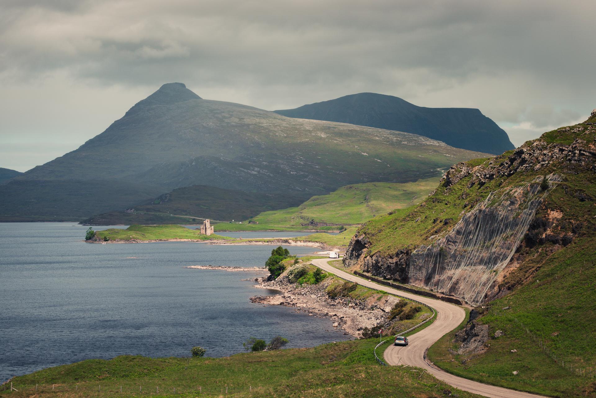 A winding road runs along a lake with green hills and rocky cliffs, leading toward a distant old stone building, with large mountains in the background under cloudy skies.