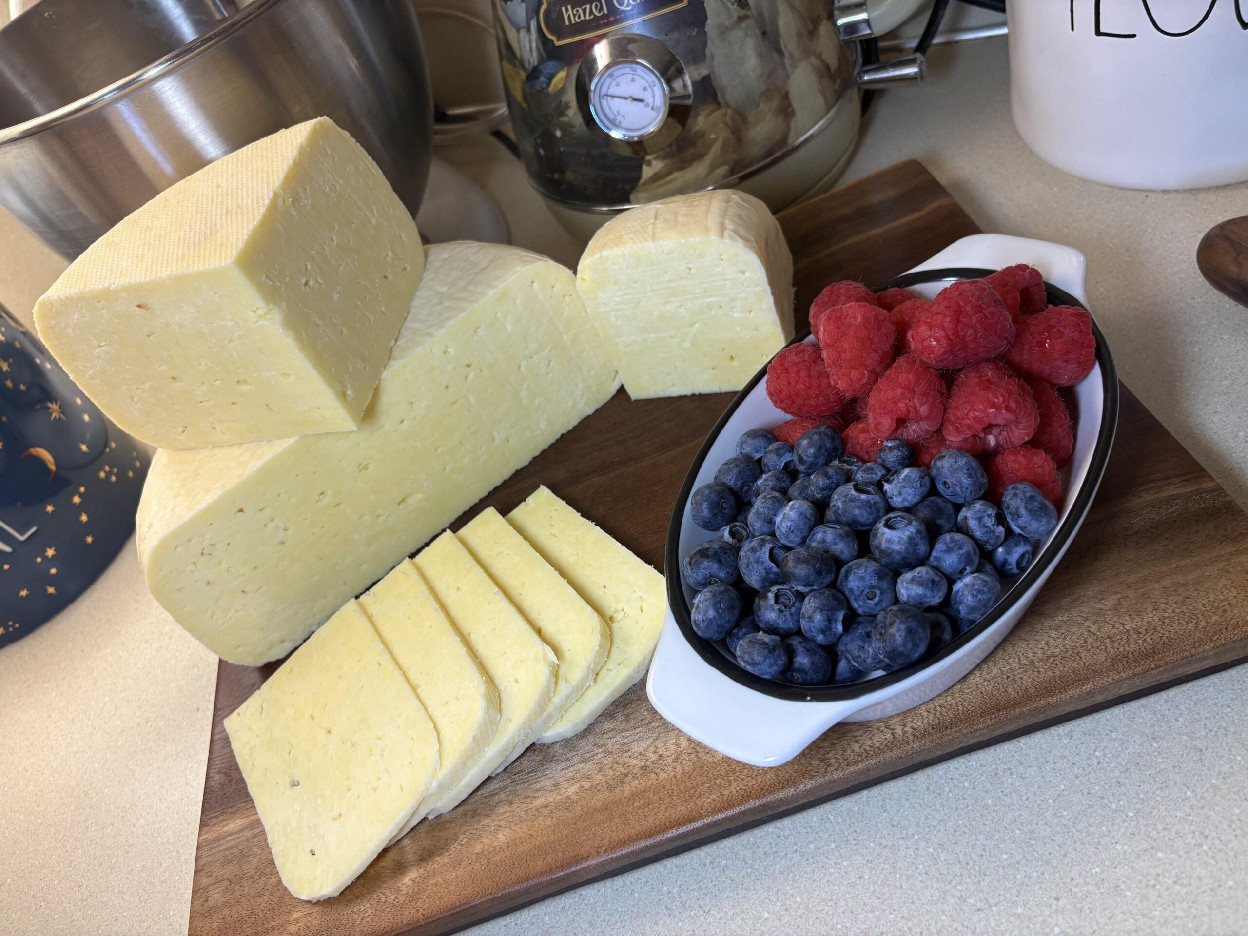 A wooden board displays blocks and slices of cheese next to a small dish filled with blueberries and raspberries, set on a kitchen counter with a mixer and other kitchen items in the background.