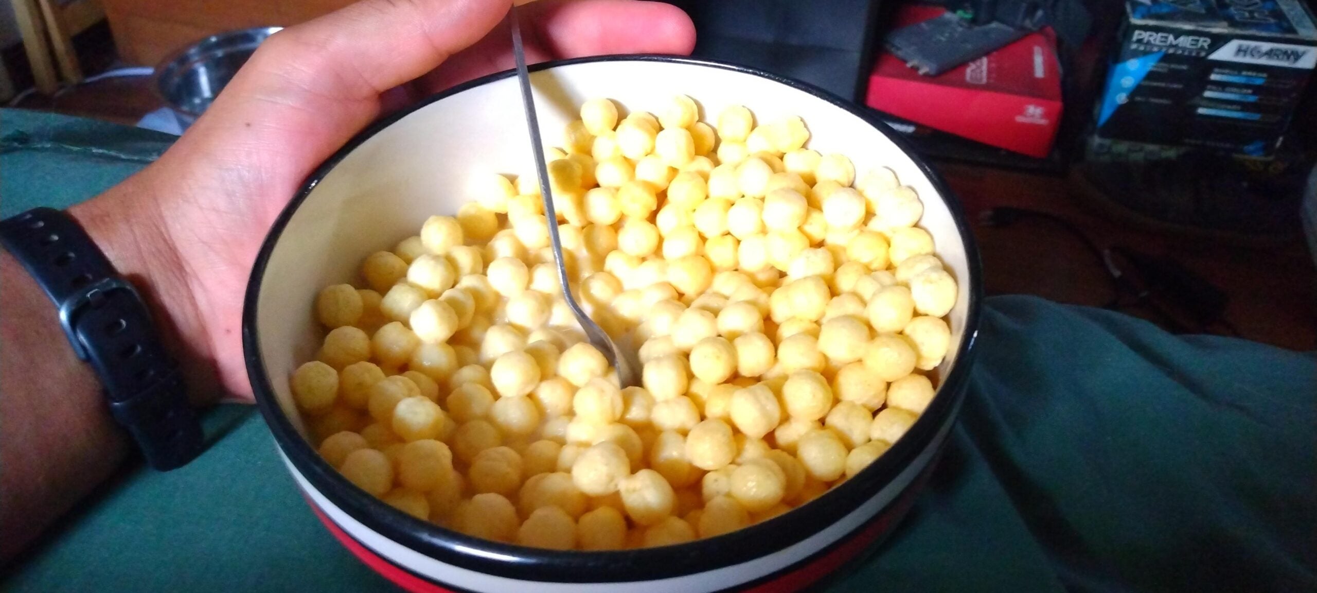 A hand holds a bowl of round yellow cereal with a spoon inside. Electronics and a green cloth appear in the blurry background.