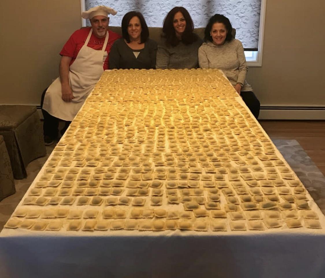 Four people sit behind a large table covered with neatly arranged homemade ravioli on a white cloth. One person wears a chef hat and apron, and all are smiling at the camera.