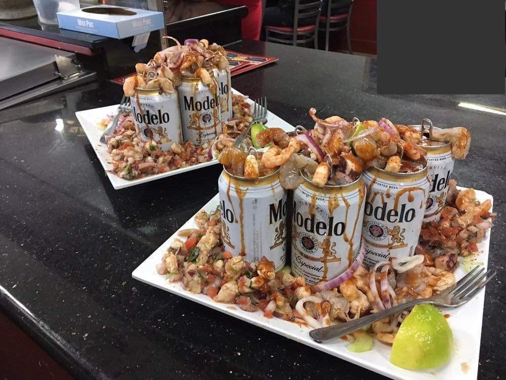 Two plates with Modelo beer cans surrounded by seafood, lime, chopped vegetables, and garnished with grilled onions and shrimp sit on a black countertop.