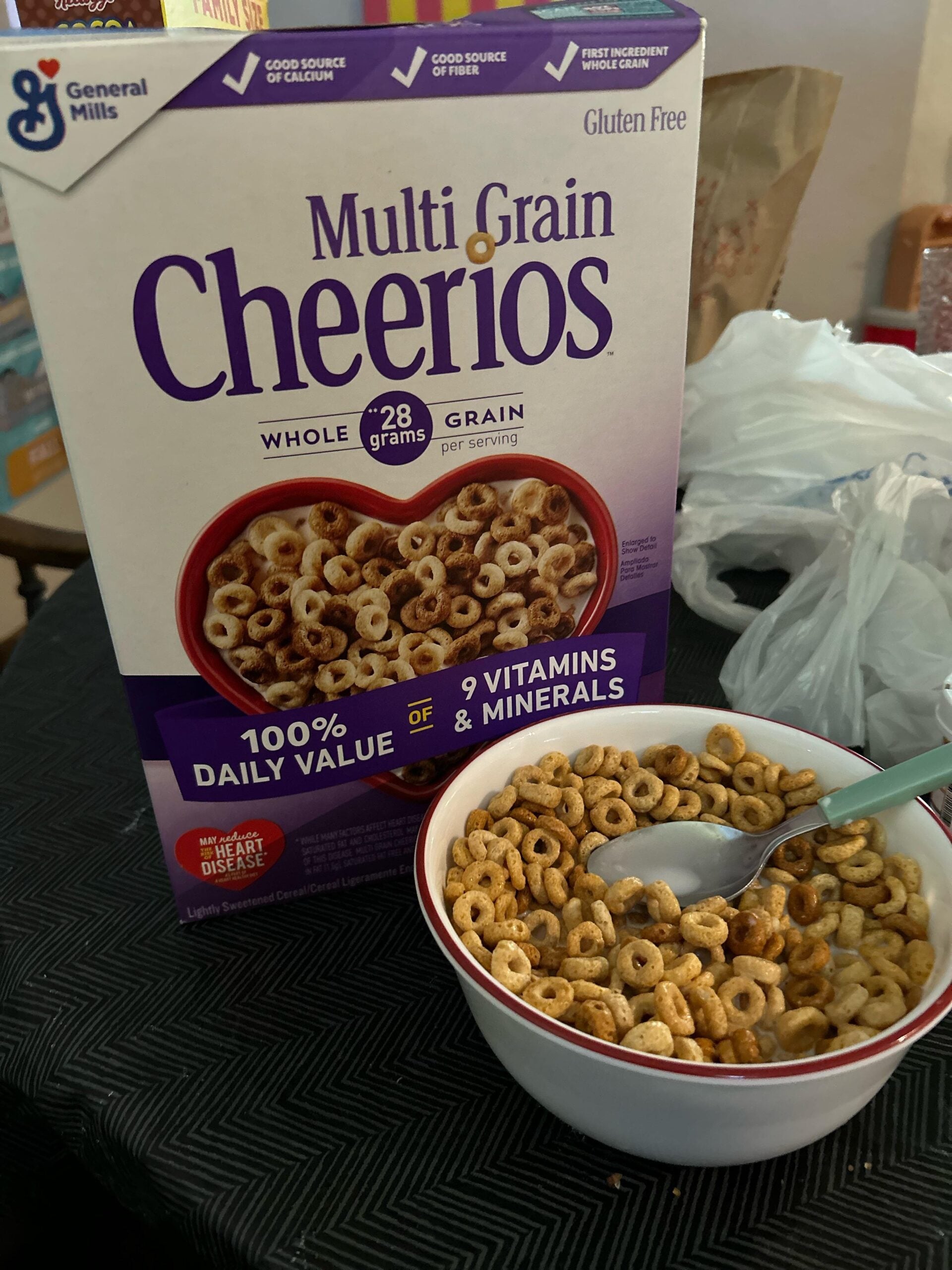 A box of Multi Grain Cheerios cereal is behind a bowl filled with the cereal and milk. A spoon rests in the bowl, which is placed on a dark textured tablecloth.