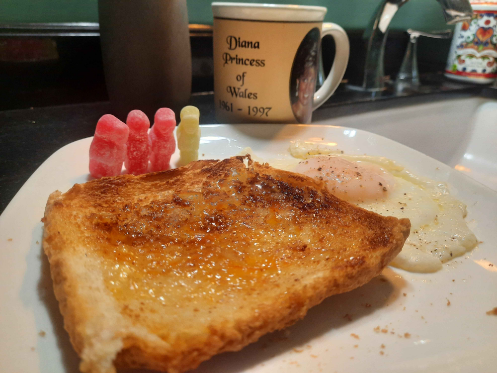 A breakfast plate with toast, two sunny-side-up eggs, three pink gummy candies, and a mug labeled “Diana Princess of Wales 1961-1997” on a kitchen counter.