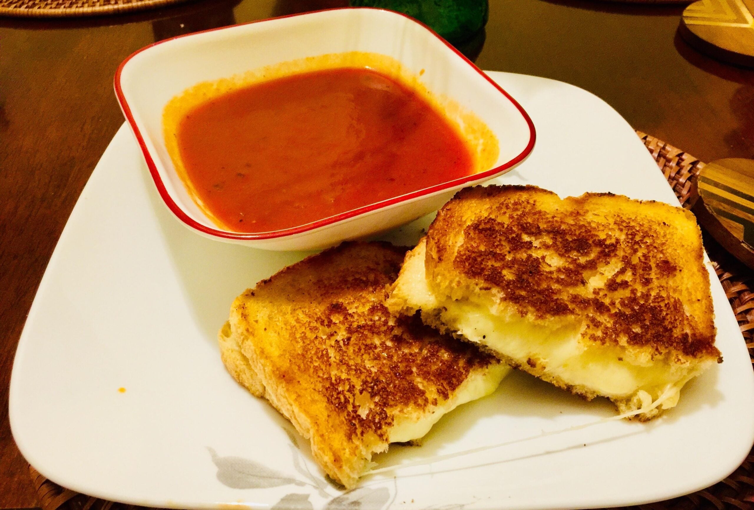 A bowl of tomato soup is served on a white plate next to two halves of golden, grilled cheese sandwiches with melted cheese, all set on a wooden table.