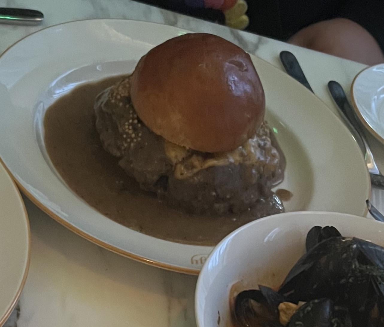 A burger with a round bun is served on a white plate, covered in brown gravy. A bowl of mussels is partially visible in the lower right corner. The table is set with cutlery.