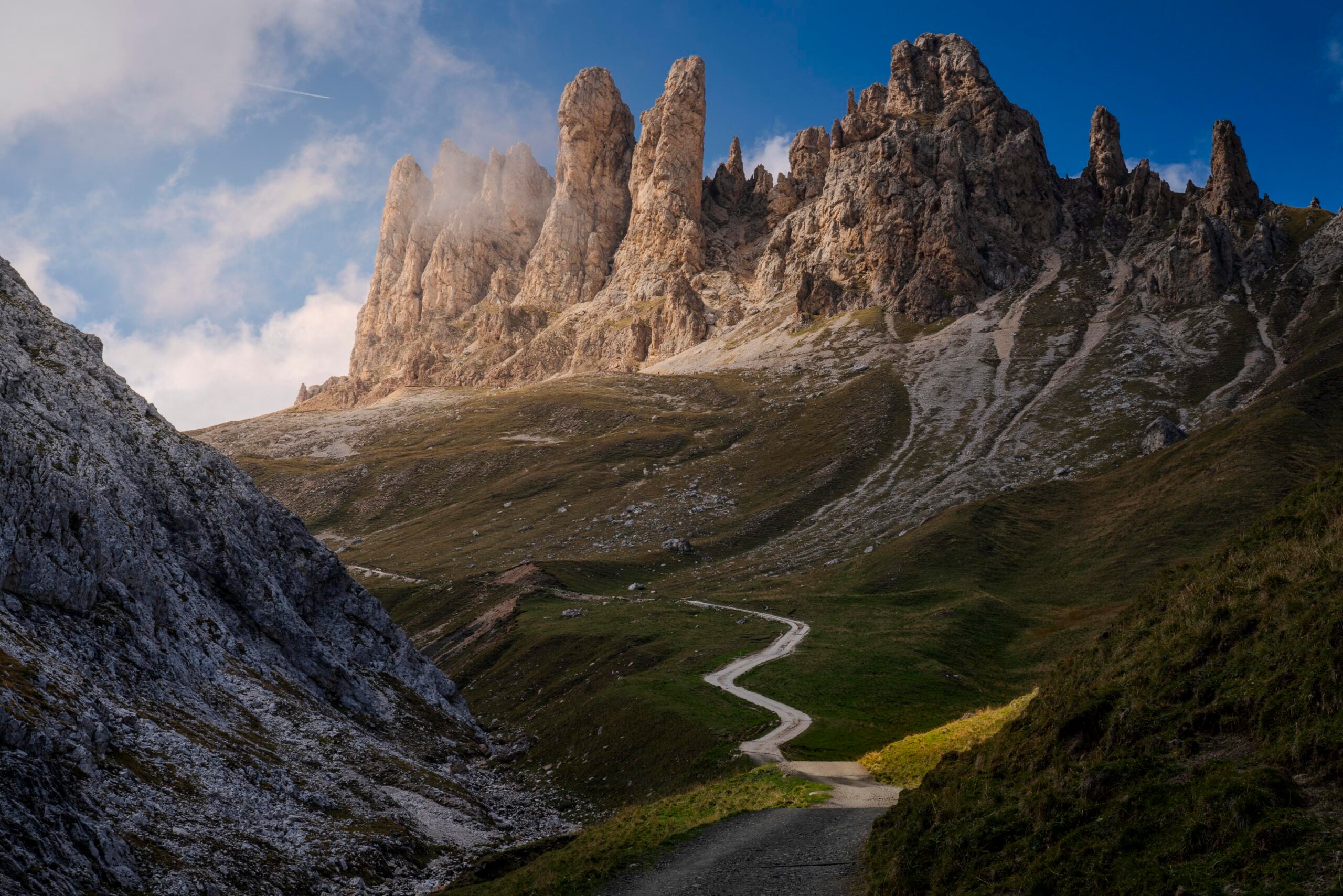 A winding dirt path leads through grassy hills toward sharp, towering mountain peaks under a blue sky with scattered clouds.