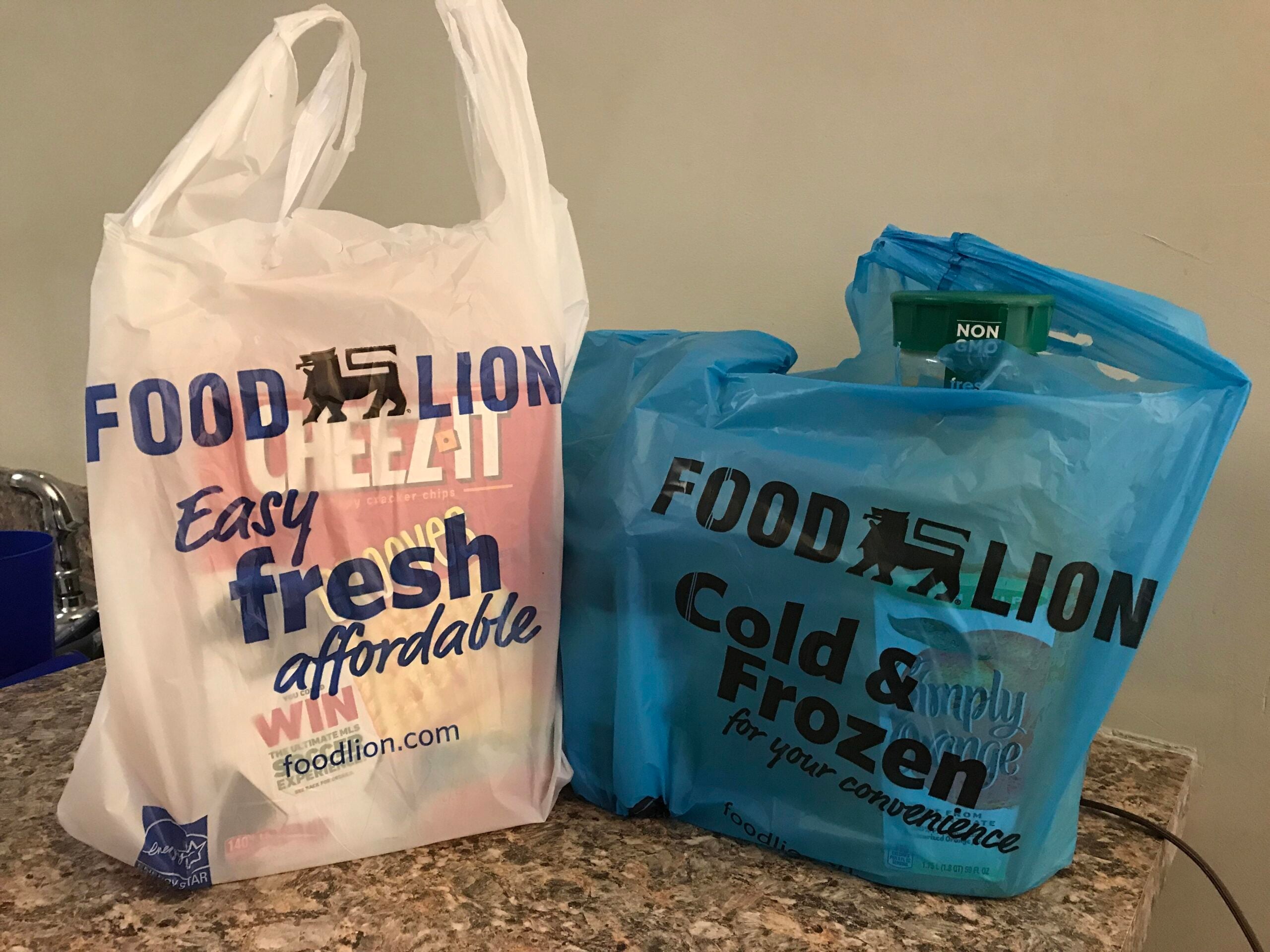 Two Food Lion grocery bags, one white and one blue, sit on a kitchen counter. The white bag holds a box of Wheat Thins, while the blue bag contains groceries, including a visible green package.