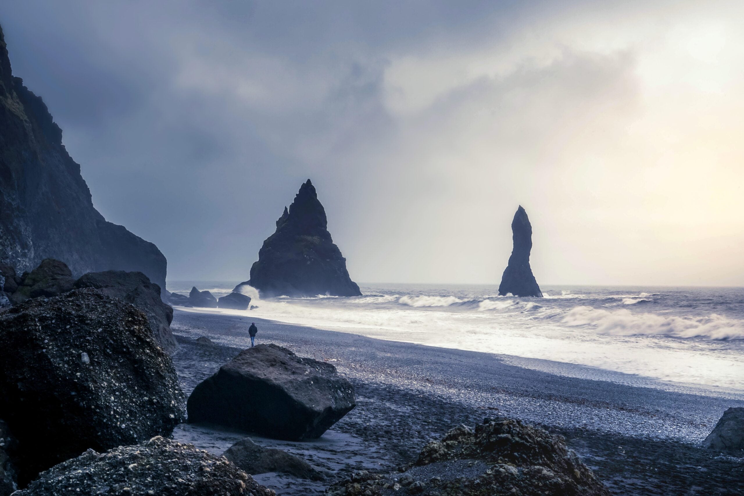 A dramatic rocky coastline with large sea stacks, dark pebbled beach, crashing waves, and misty skies; a lone person stands near a boulder, gazing at the ocean along one of the region’s incredible beaches.
