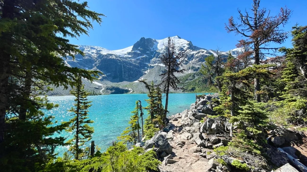 A turquoise lake surrounded by rocky terrain and evergreen trees, with a snow-capped mountain in the background under a clear blue sky.