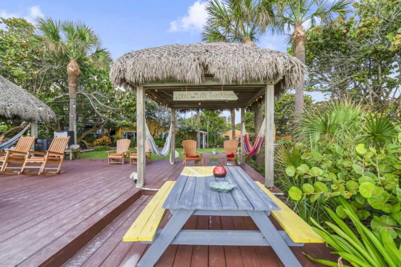 A wooden picnic table with yellow benches sits in front of a tiki hut with hammocks, Adirondack chairs, and palm trees, in a tropical garden setting under a blue sky—evoking the relaxed atmosphere found at many resorts.