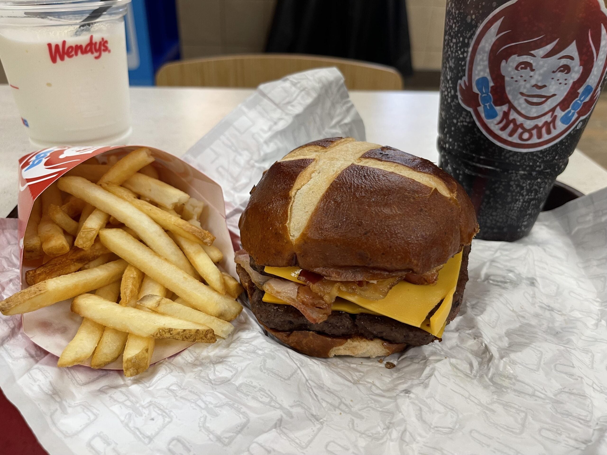 Wendy’s meal with a cheeseburger on a pretzel bun, bacon, and cheddar cheese, served with a side of fries, a soft drink in a Wendy’s cup, and a vanilla Frosty on a table.
