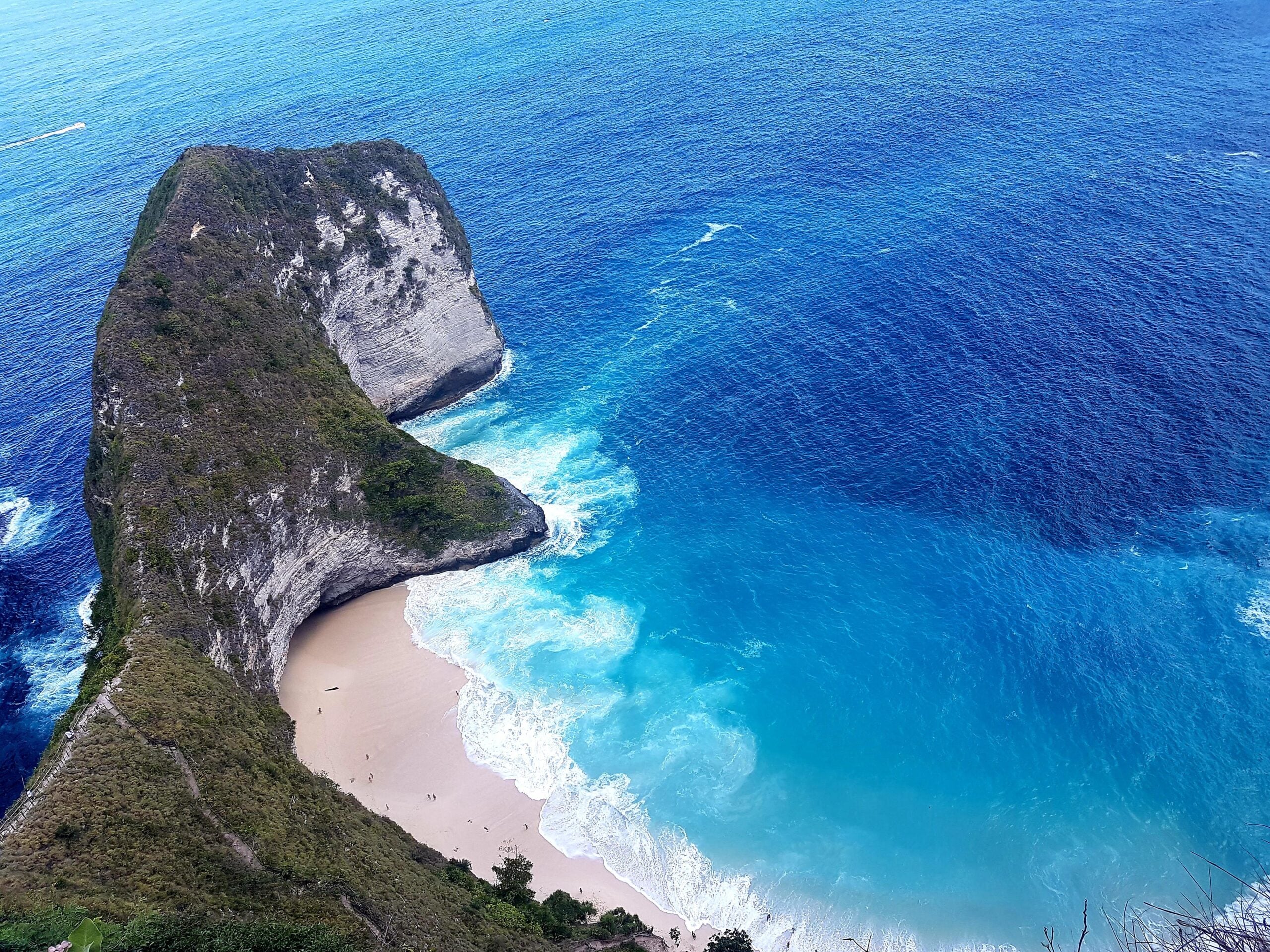 Cliffside view of a curved, rocky peninsula with lush greenery, surrounded by bright blue ocean, and a small sandy beach with gentle waves below.