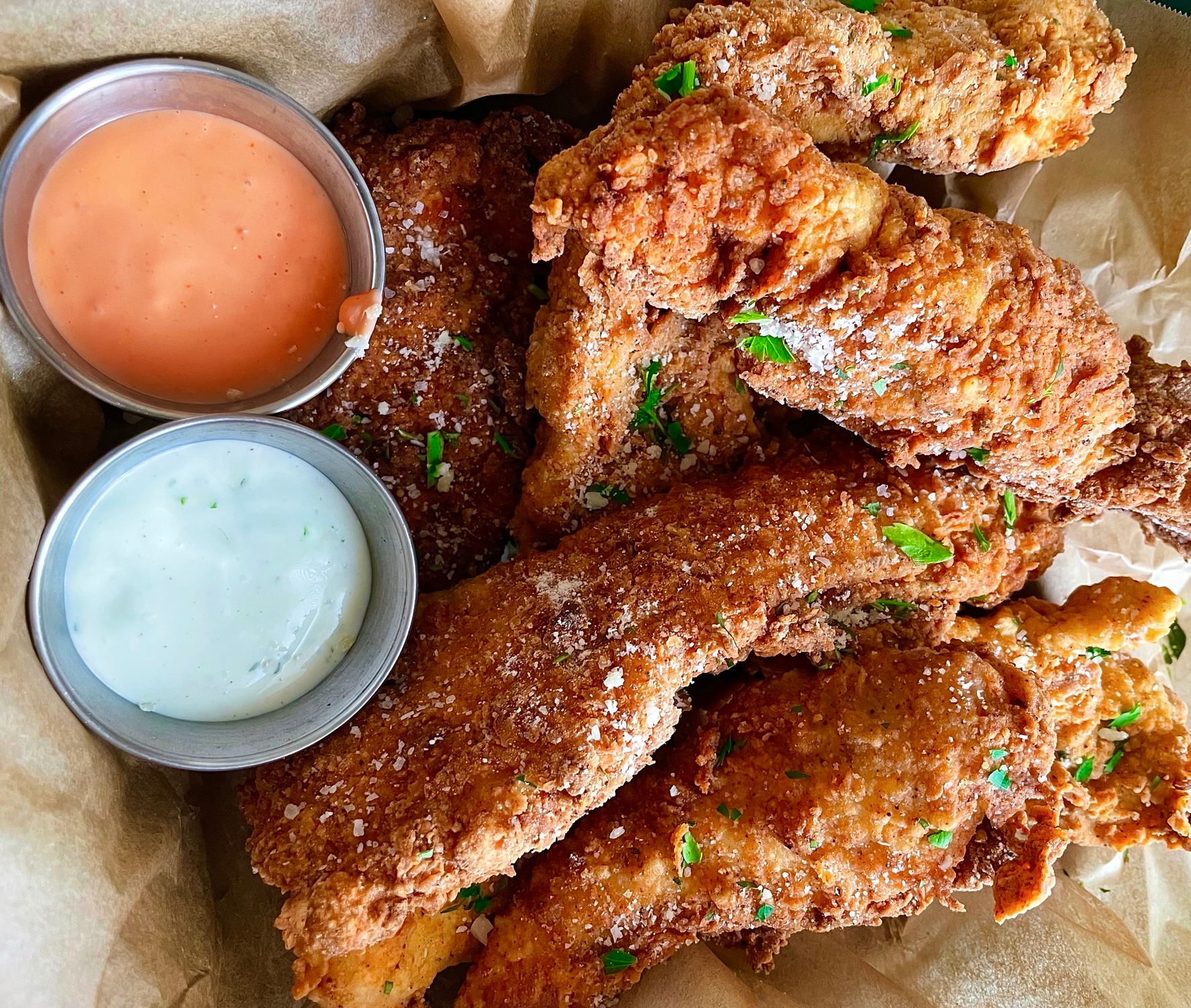 A basket of crispy fried chicken tenders garnished with herbs, served with two dipping sauces—one creamy white and one orange—in small metal cups.