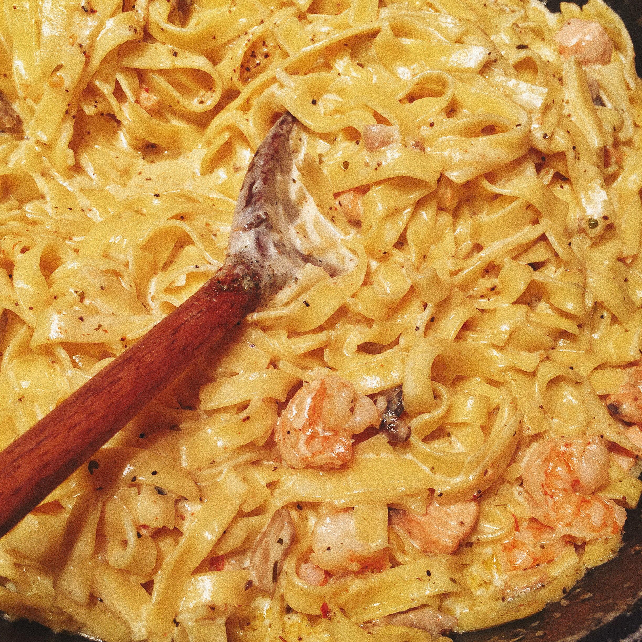 Tagliatelle pasta mixed with shrimp in a creamy sauce, topped with black pepper. A wooden spoon coated with sauce rests in the pasta, suggesting the dish has just been stirred.