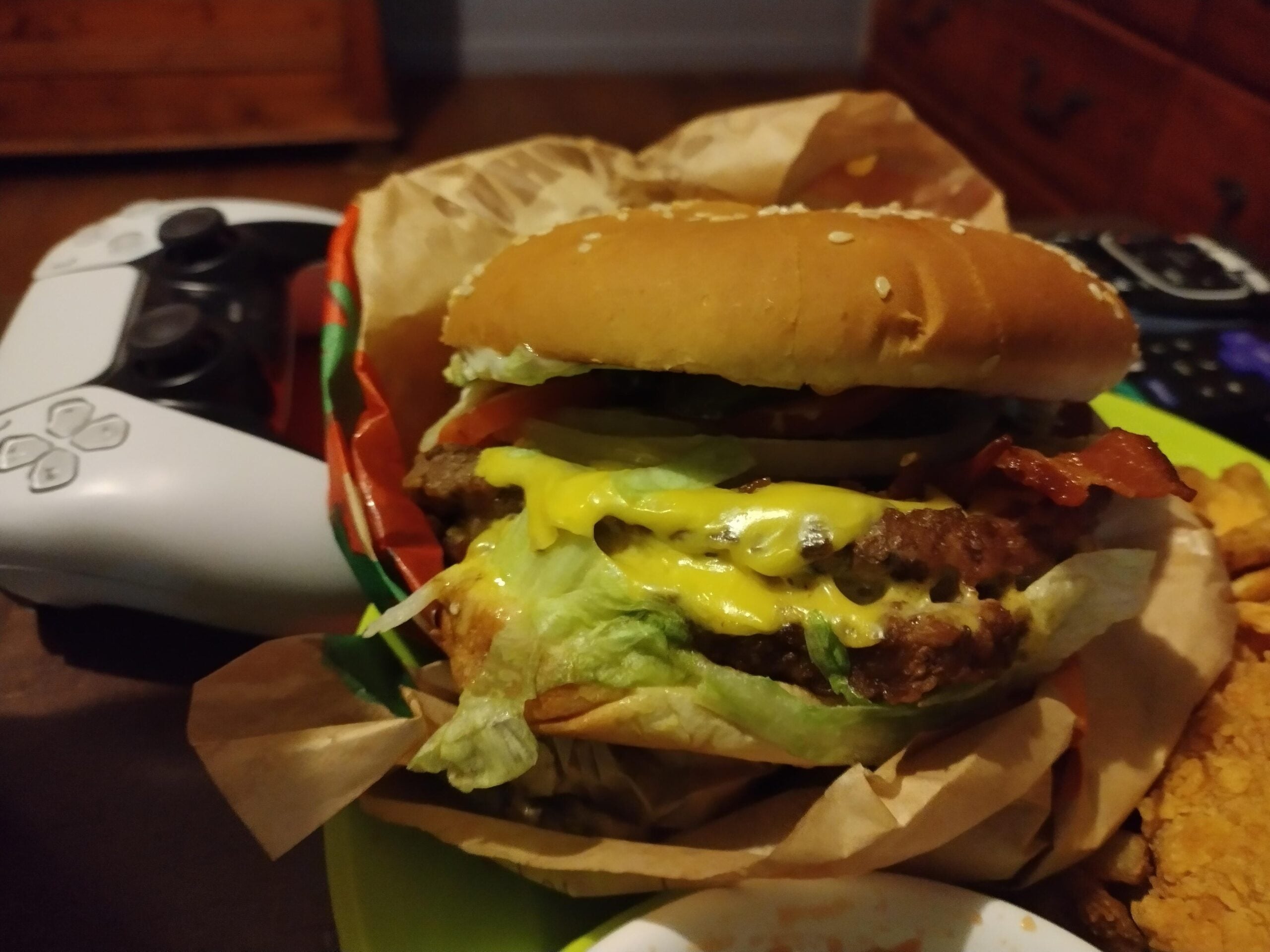 A close-up of a cheeseburger with lettuce, tomato, bacon, and melted cheese, partially wrapped in paper. A video game controller sits in the background on a table next to the burger.