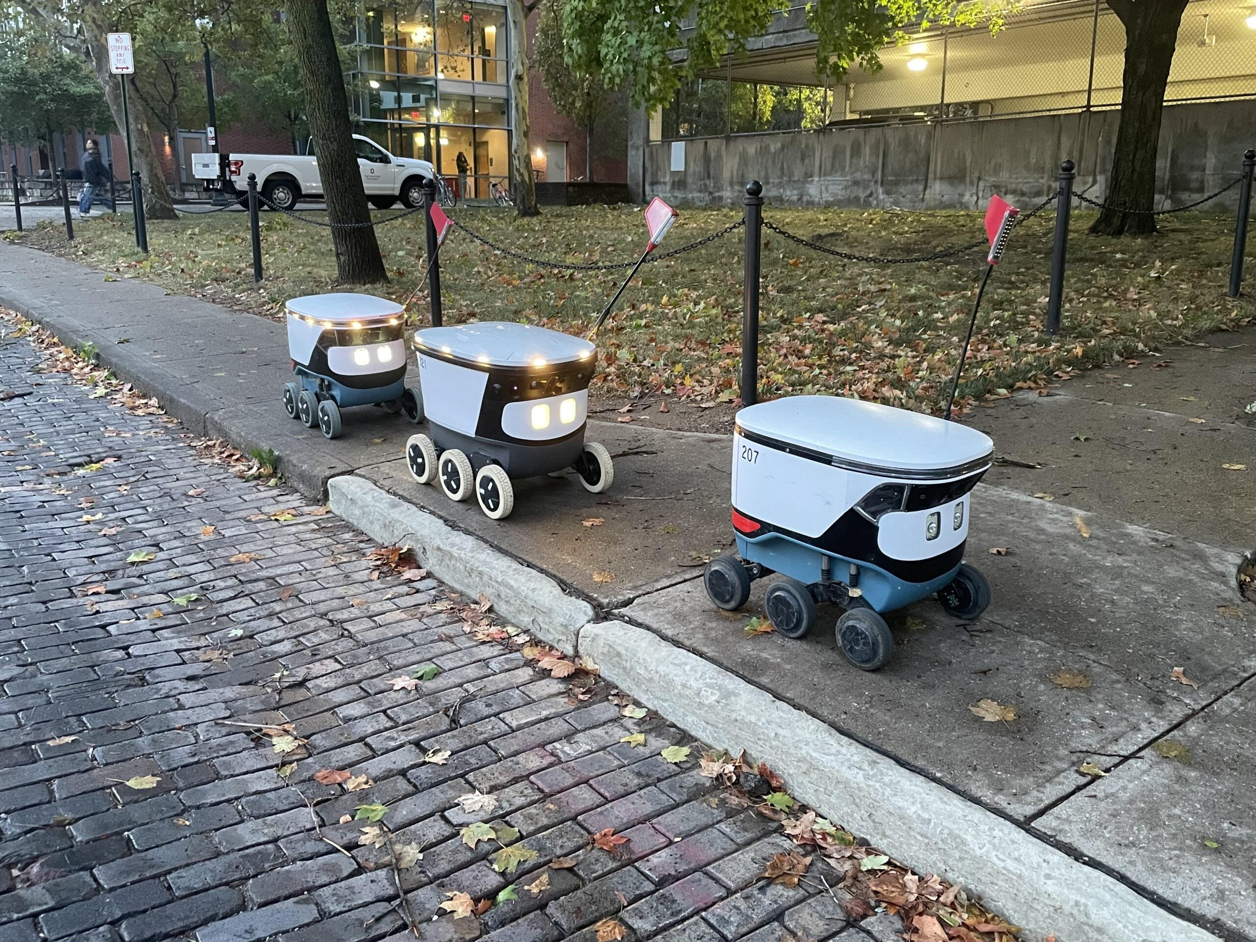 Three small delivery robots with headlights and flags wait on a sidewalk curb next to a street with fallen leaves. Trees, a parked car, and a building are in the background.