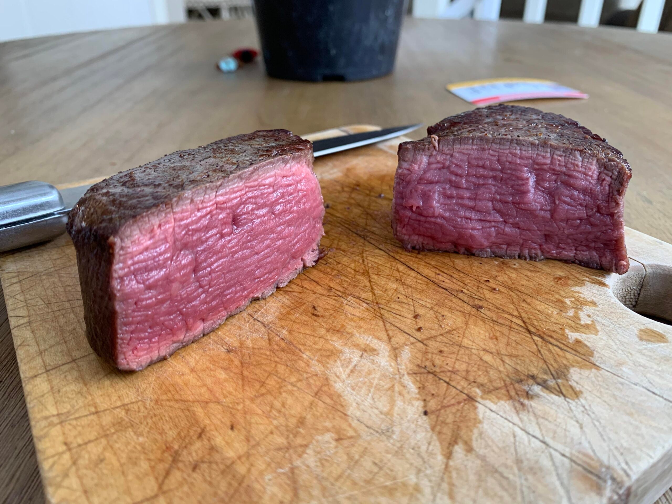 Two thick slices of rare-cooked beef steak resting on a wooden cutting board, showing a seared brown exterior and a pink, juicy interior. A knife and some blurred objects are visible in the background.