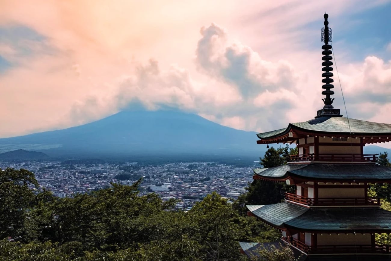 A traditional Japanese pagoda stands in the foreground with Mount Fuji and a cityscape—one of the Biggest Cities In The World—in the background under a colorful, cloud-filled sky.