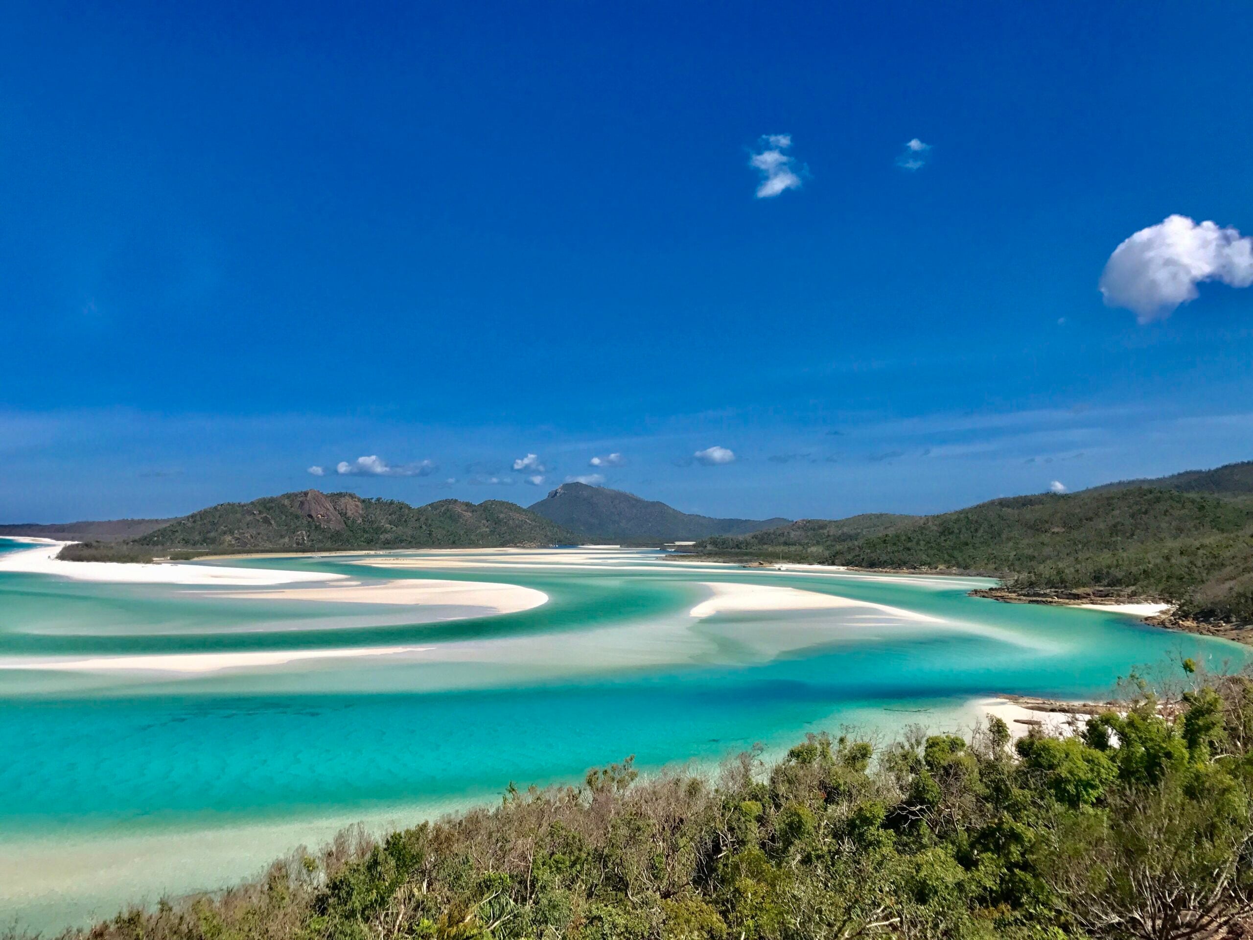 Turquoise water with white sandbars curves along the shore, surrounded by green hills under a clear blue sky with a few clouds—one of the most incredible beaches you’ll ever discover.